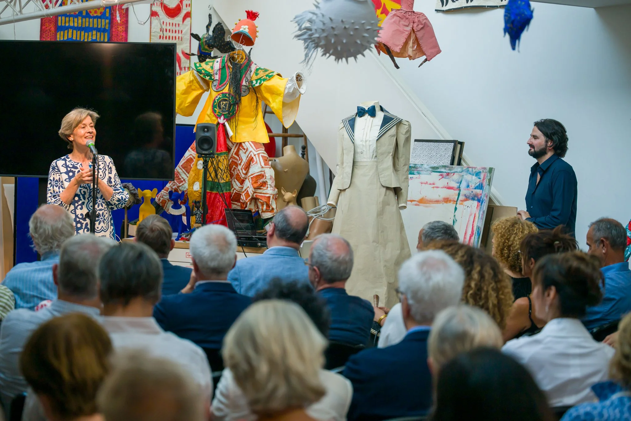Une femme parle devant un public lors d'une présentation ou d'une conférence, avec diverses œuvres d'art et une sculpture colorée en arrière-plan.