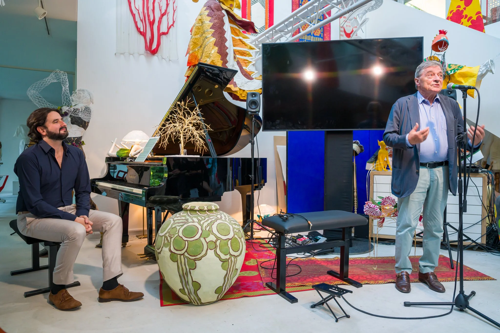Un homme âgé parle au microphone lors d'une performance musicale, un pianiste l'écoute assis à côté d'un piano noir, dans un espace décoré artistiquement avec un grand écran, des œuvres d'art murales, et des meubles colorés.