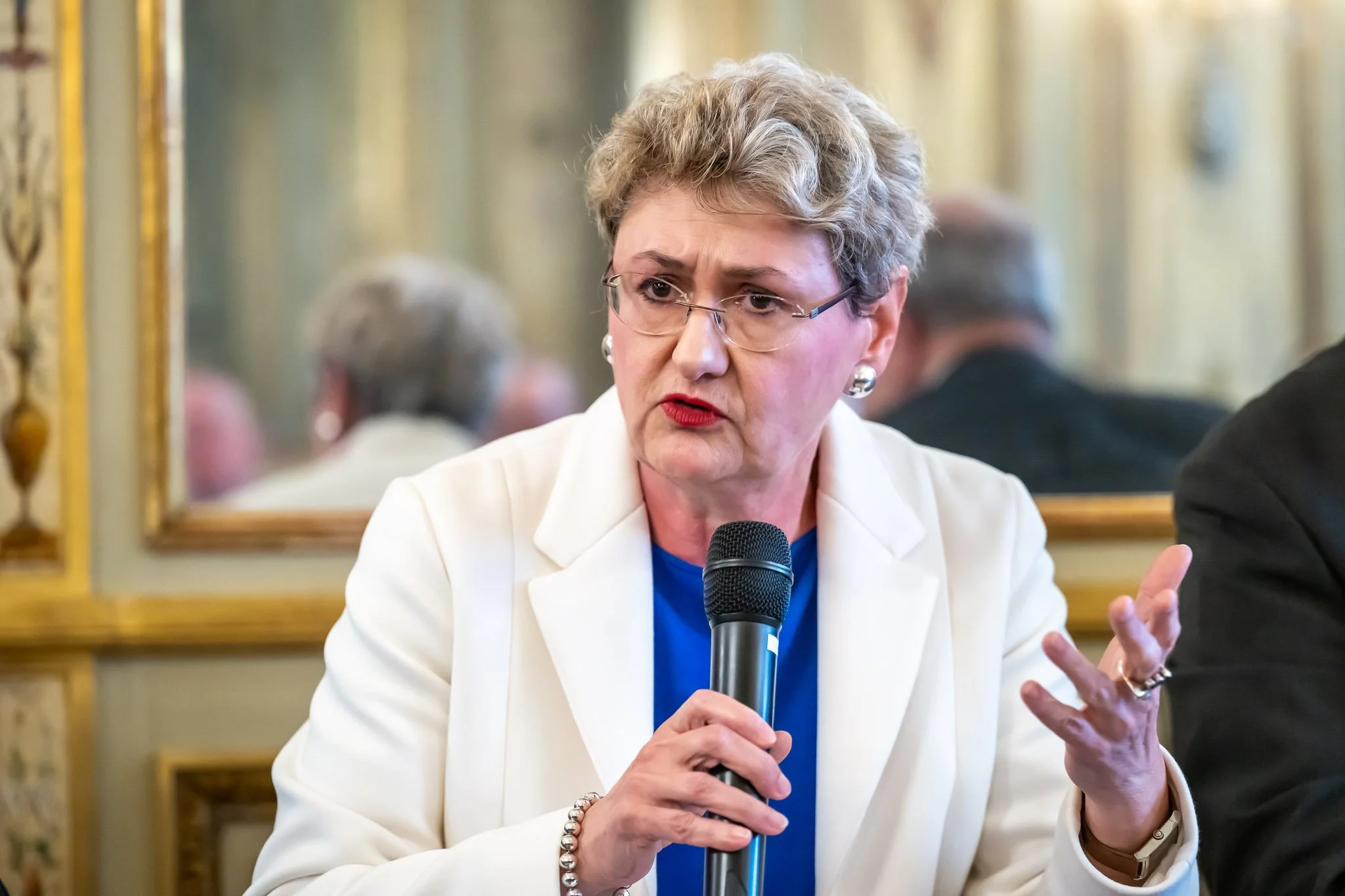 Woman holding a microphone, wearing a white suit, speaking at an event.