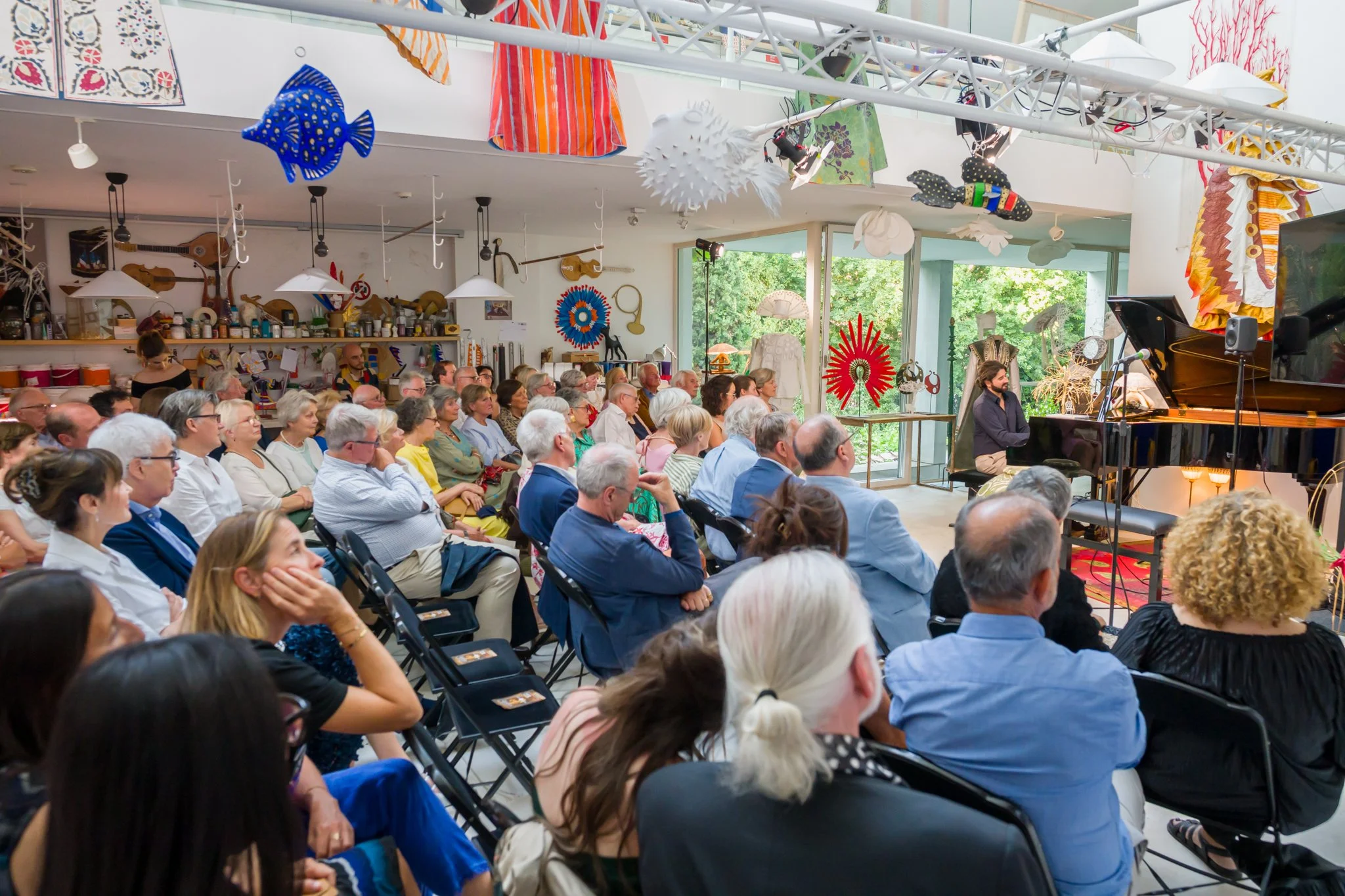 Public attentif lors d'un concert de piano dans une salle décorée avec des œuvres d'art suspendues et des objets d'artisanat colorés.