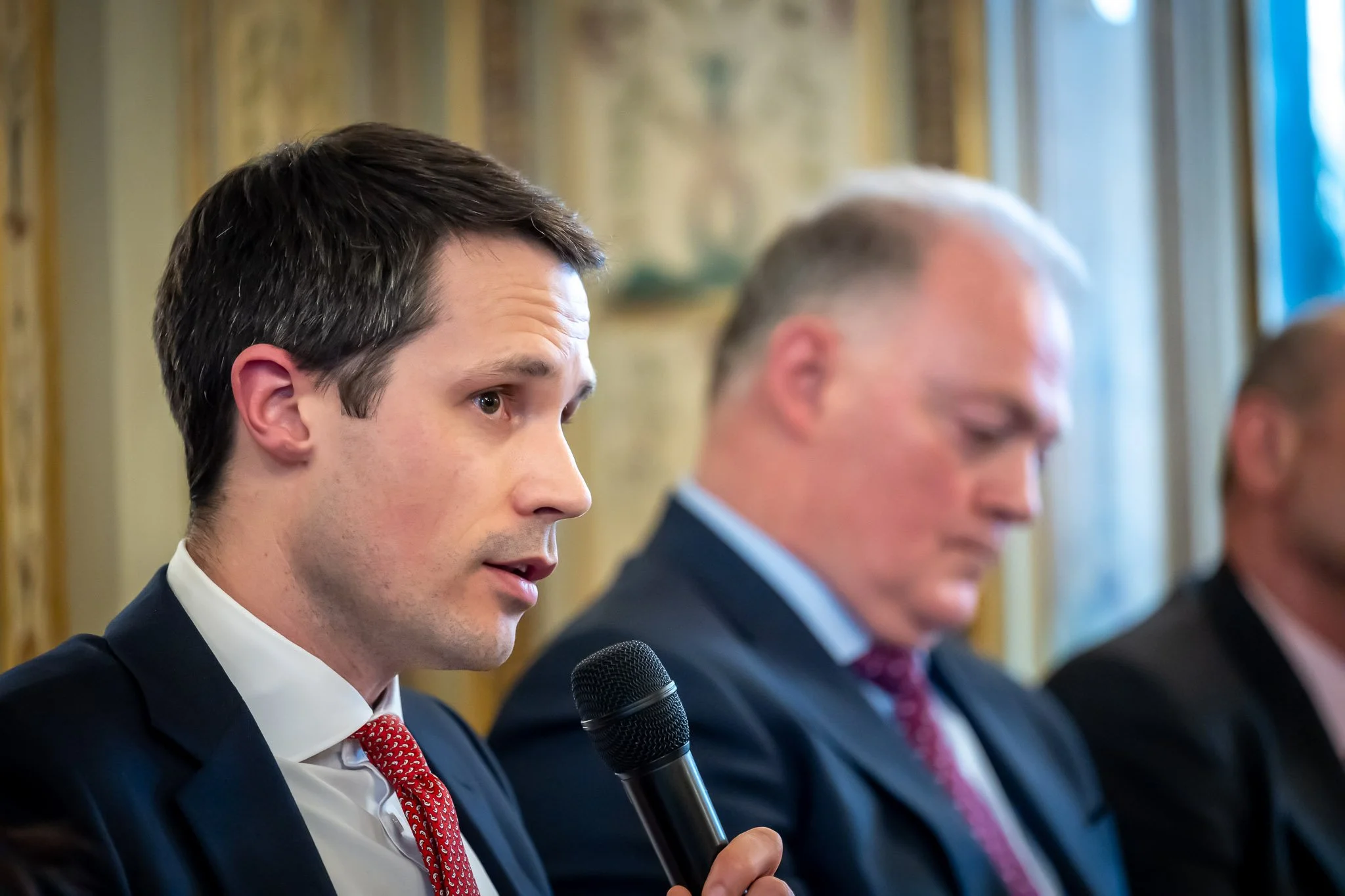 A man in a suit speaks into a microphone at a conference, with another man in a suit in the background.