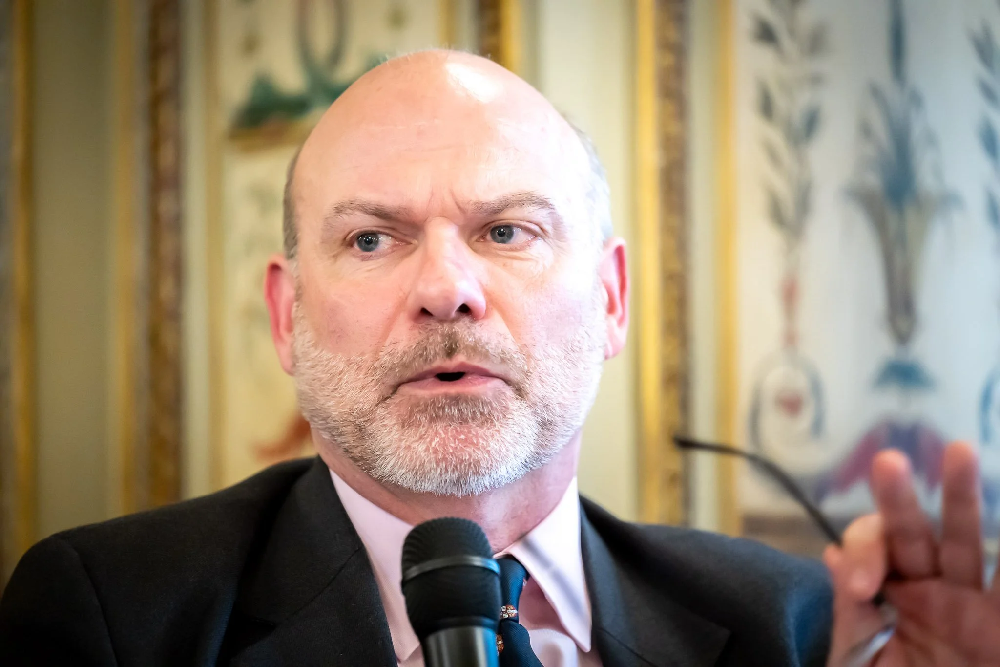 Man speaking into a microphone with serious expression, holding glasses, wall decor in background.