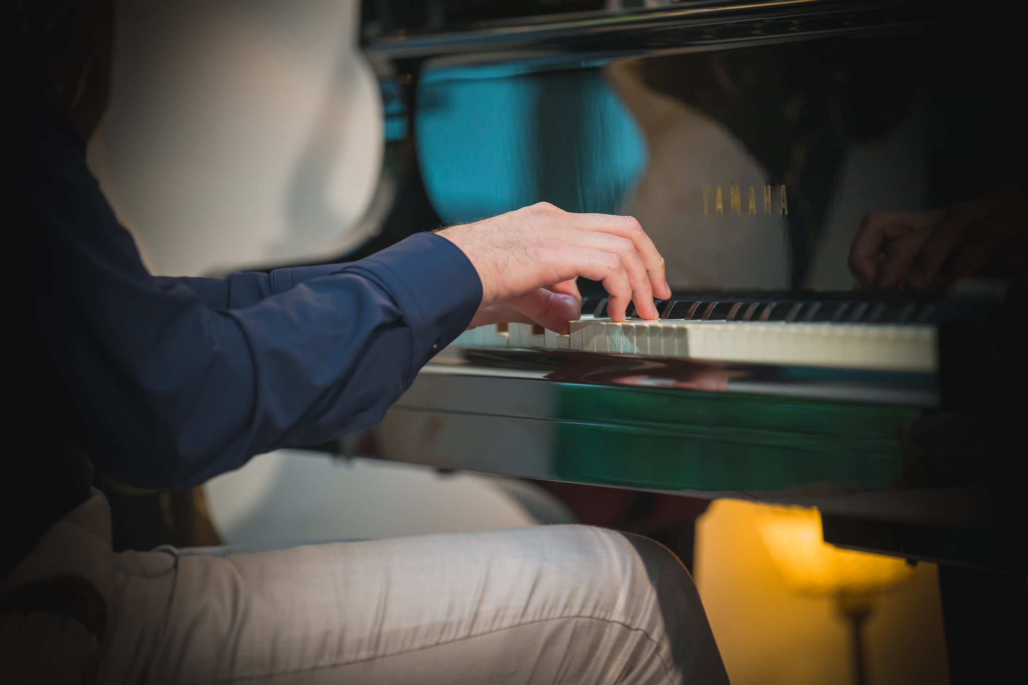 Personne jouant du piano noir à queue, portant une chemise bleu et un pantalon beige, dans un intérieur.