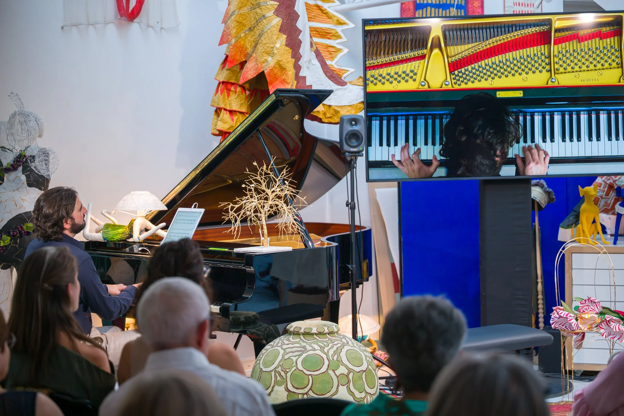 Un musicien joue du piano lors d'une performance ou d'une présentation devant un public, avec un écran vidéo montrant un autre pianiste. La scène est décorée avec des objets artistiques colorés et des aménagements décoratifs.