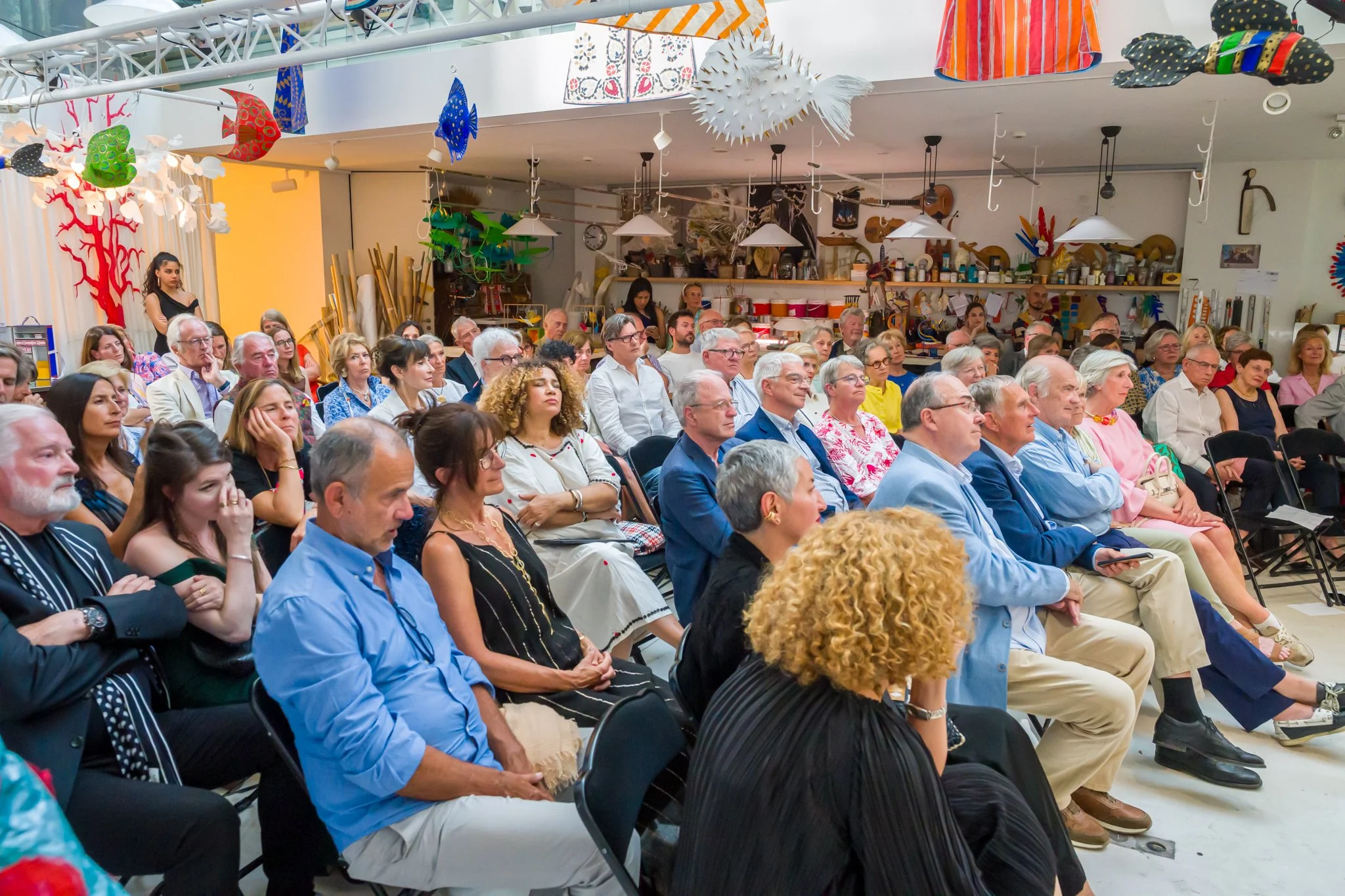 Audience de personnes assises dans une salle, écoutant attentivement lors d'une présentation ou conférence, avec des décorations colorées suspendues au plafond.