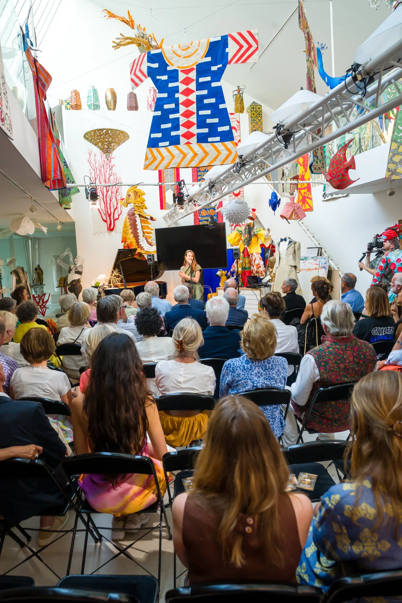 Une salle d'exposition avec des spectateurs regardant un spectacle ou une conférence. Le plafond est décoré de tissus colorés et d'œuvres d'art suspendues, avec un piano sur scène et une femme parlant à l'audience.