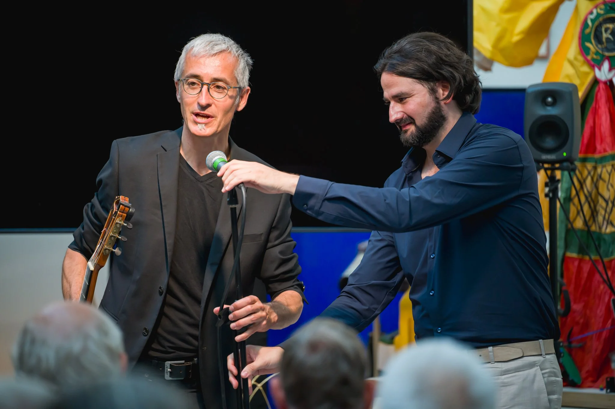 Deux hommes parlant devant un public lors d'un événement, avec un microphone, l'un d'eux tenant une guitare, en arrière-plan un écran noir et des décorations colorées.
