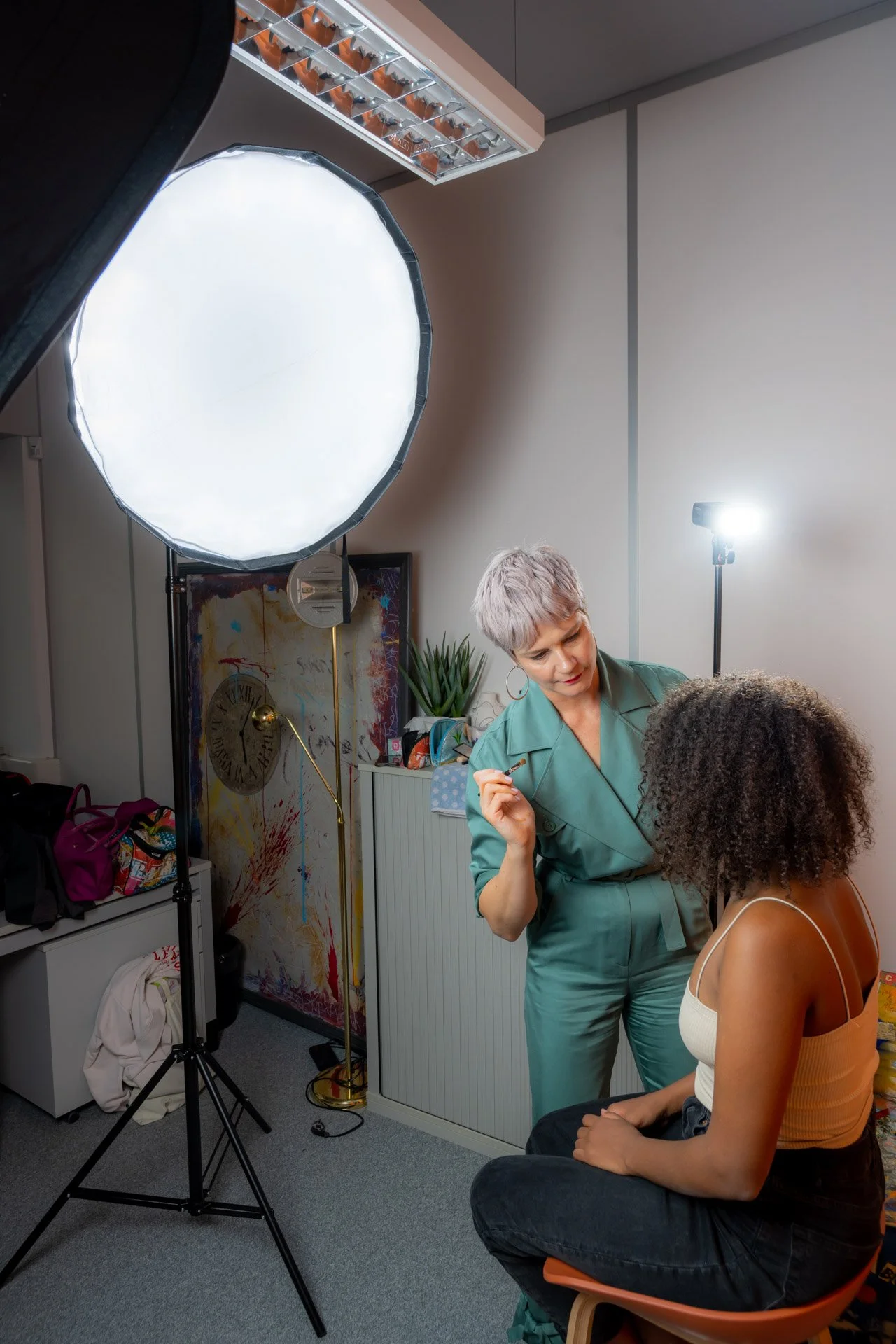 Une fixatrice de maquillage travaille sur une femme assise dans un studio avec un éclairage professionnel et des accessoires de photographie.