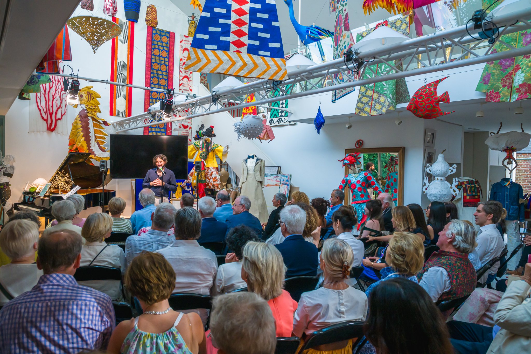 Une salle d'exposition vibrant avec des œuvres d'art colorées, décorations suspendues, mannequins habillés de vêtements colorés, et un public assis écoutant une personne qui parle sur scène.