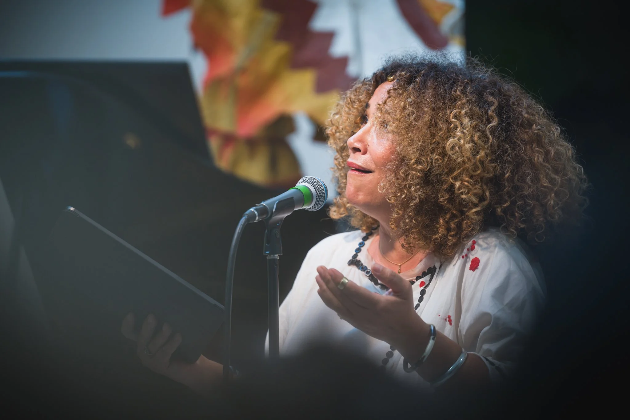 Une femme avec des cheveux bouclés, chantant ou récitant avec expression, tenant un livre ou une tablette devant un microphone lors d'une performance ou d'un discours.