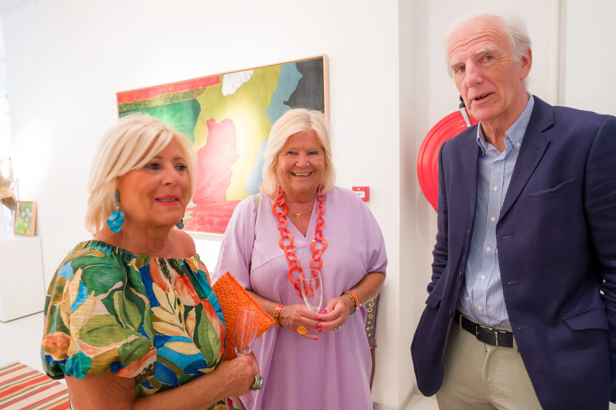 Trois personnes souriantes lors d'une exposition d'art dans une galerie, discutant et tenant des verres en cristal.