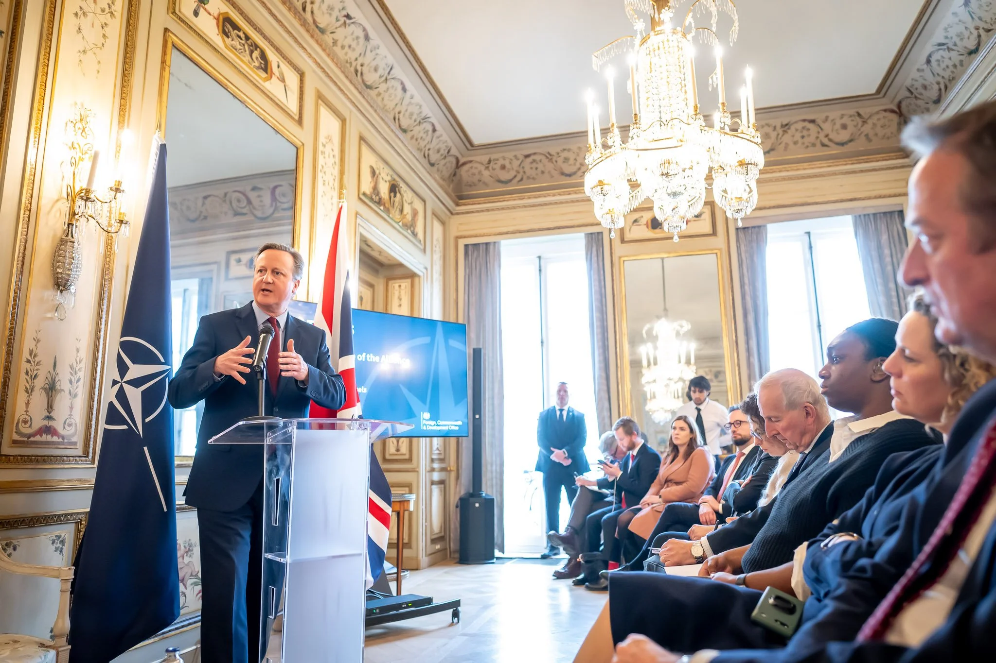 Man in suit speaking to podium at conference in elegant room with chandelier, British flag and NATO flag, and seated audience listening.