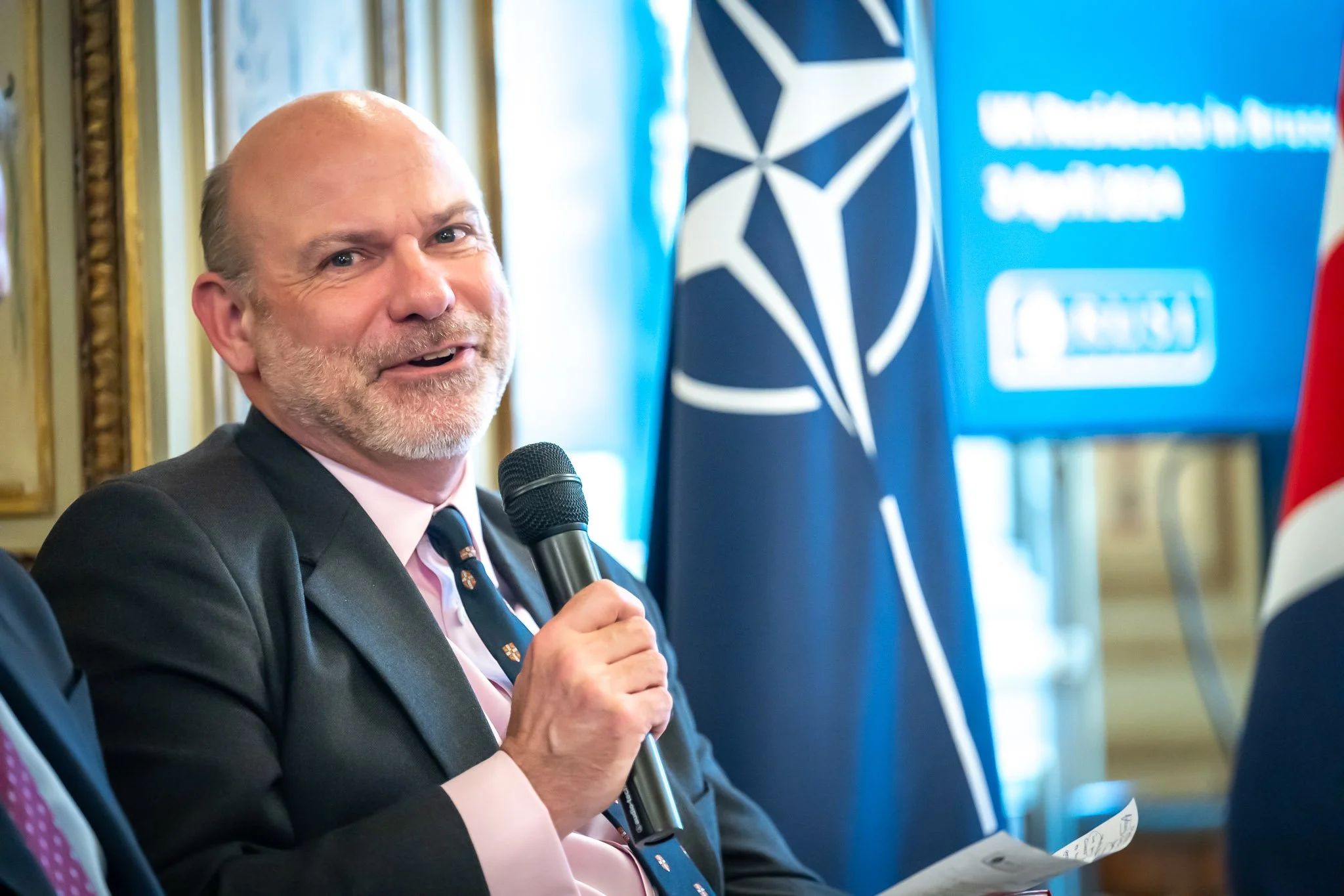 A person holding a microphone and smiling, with a NATO flag in the background.