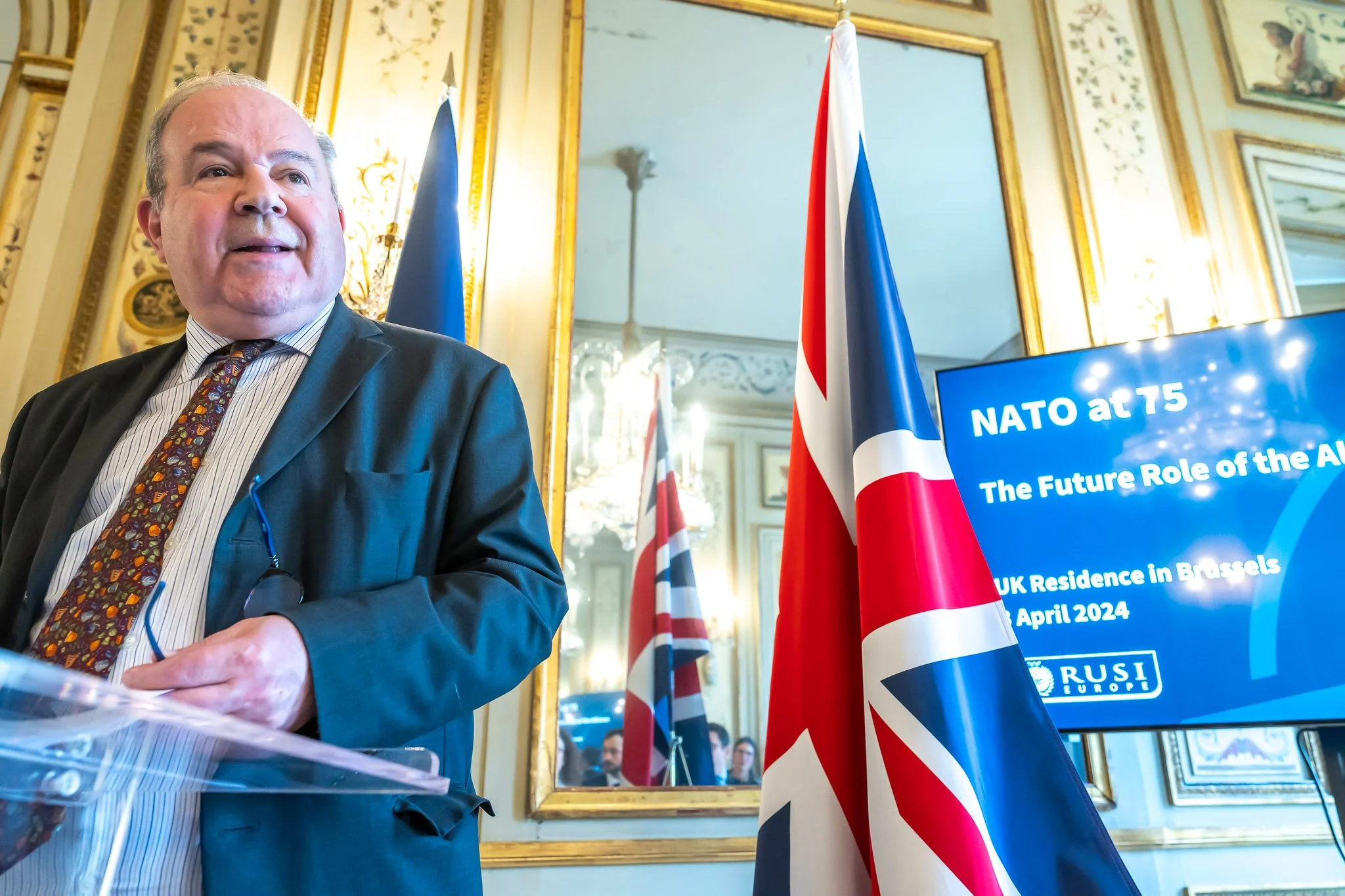 Man in suit speaking at a NATO event in Brussels, with a British flag and a screen displaying "NATO at 75".