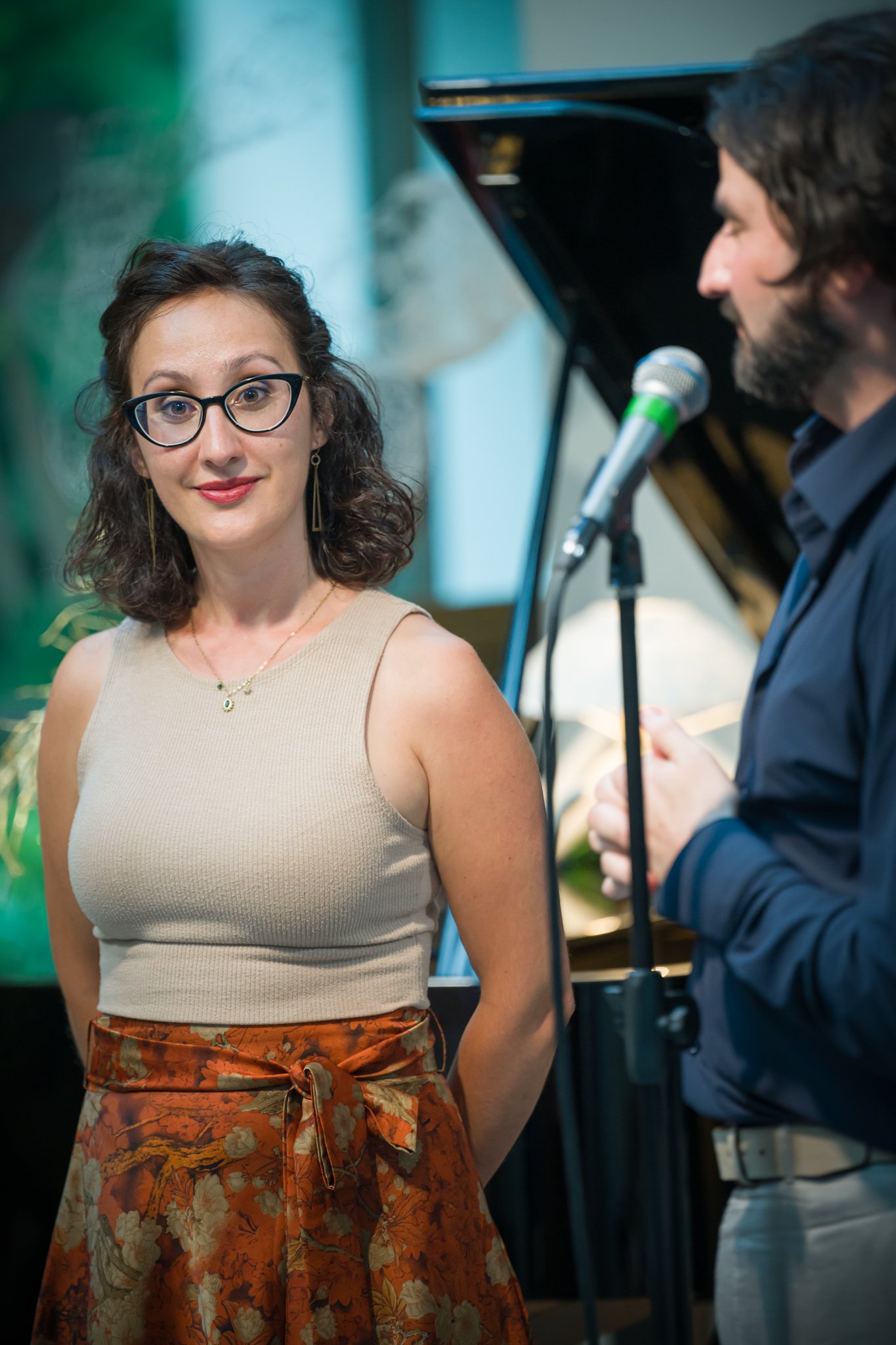 Une femme portant un haut beige et une jupe à motifs floraux est en train de regarder un homme qui parle dans un micro, avec un piano en arrière-plan.