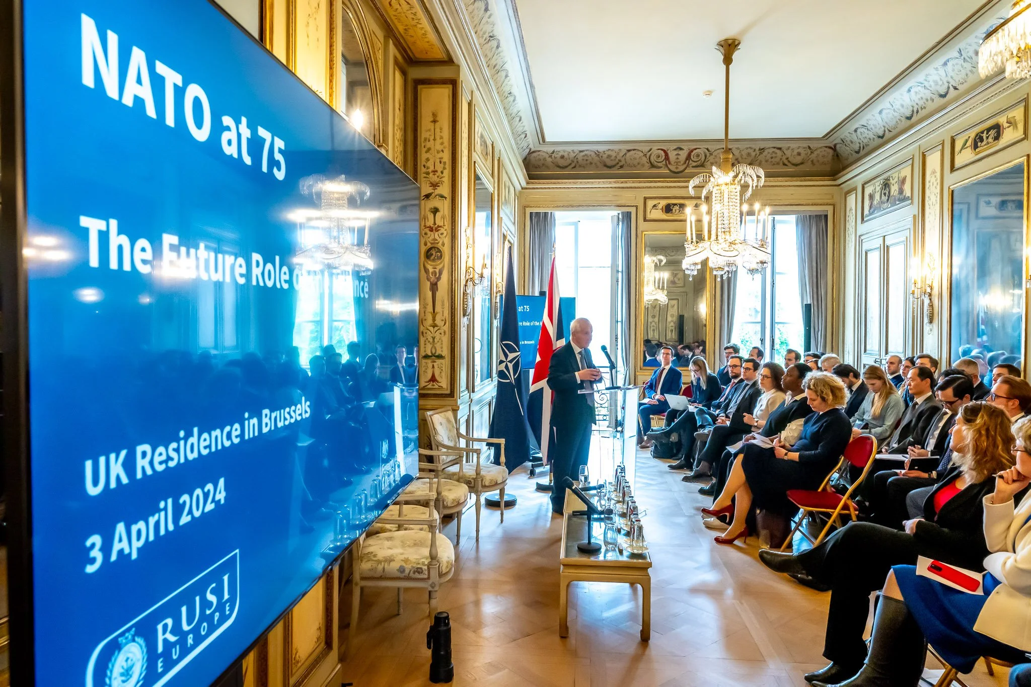 NATO 75th anniversary conference at the UK residence in Brussels, with a speaker in front of a seated audience. A large screen displays the date April 3, 2024. Elegant interior decor with chandeliers and mirrors.