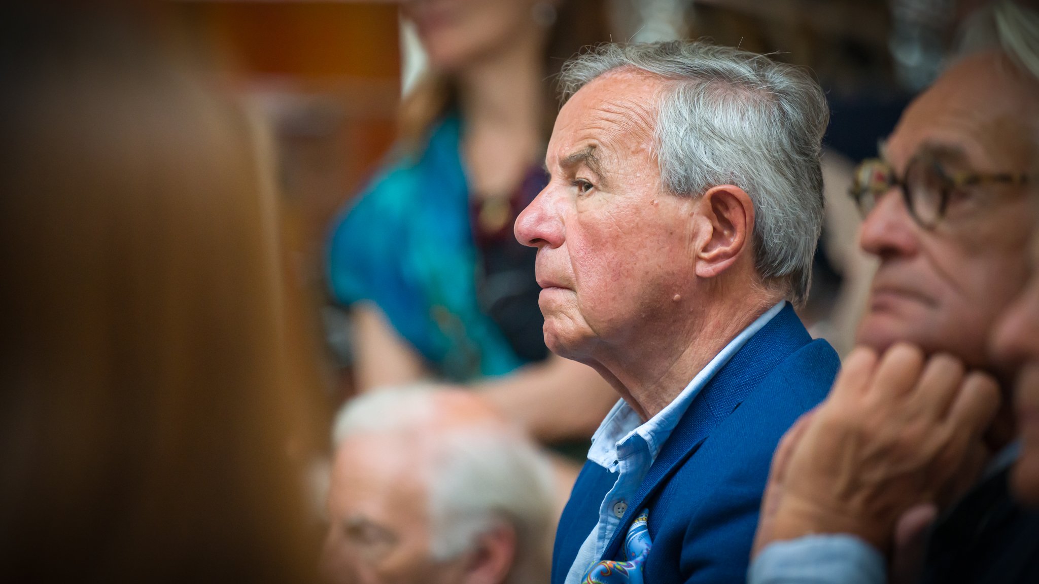 Un homme âgé en bleu assis dans une salle, écoutant attentivement lors d'une réunion ou d'une conférence.