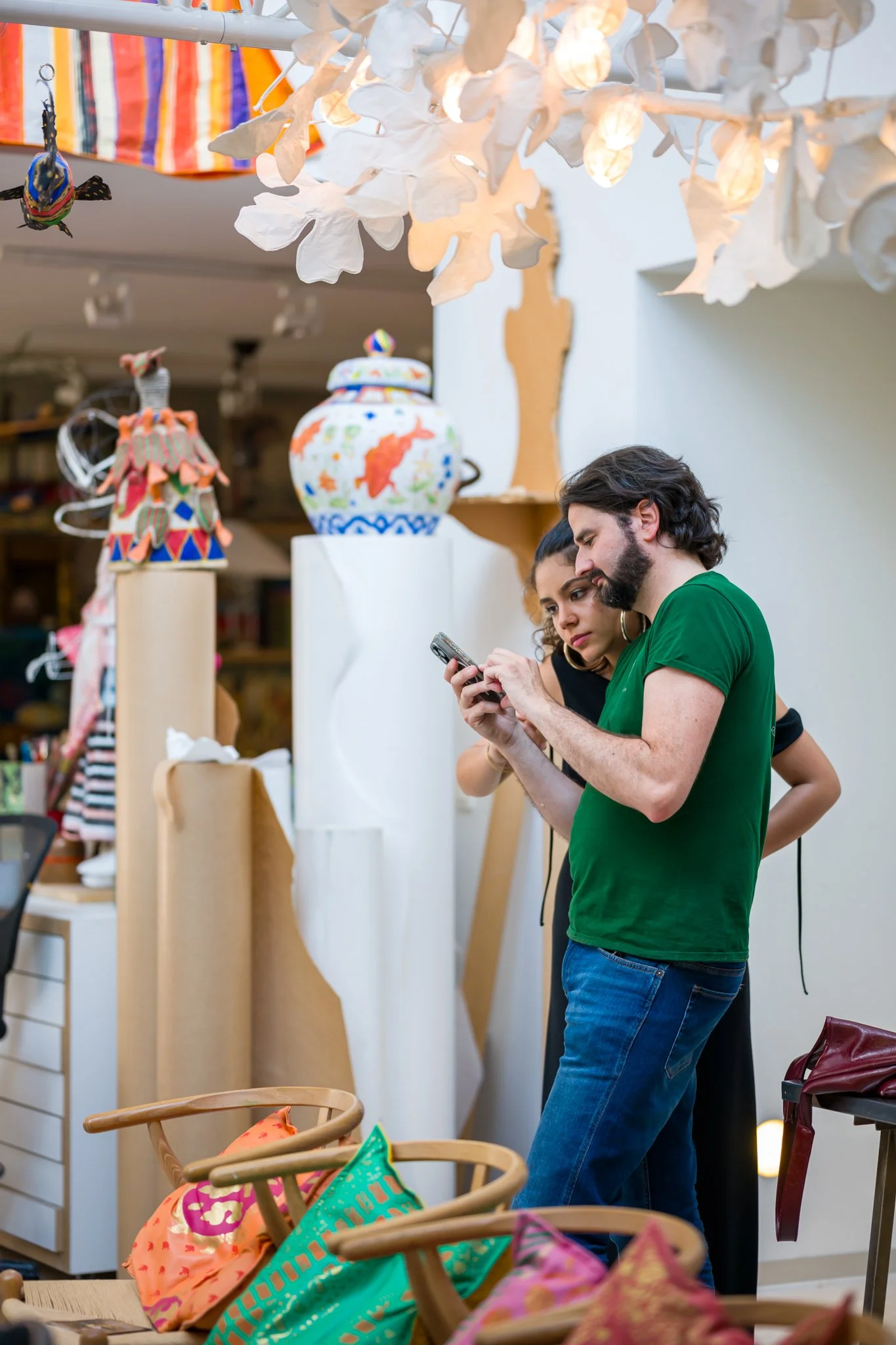 Deux personnes regardent un téléphone dans un espace d'exposition d'art ou artisanat, entourées de vêtements, tissus colorés et objets décoratifs.