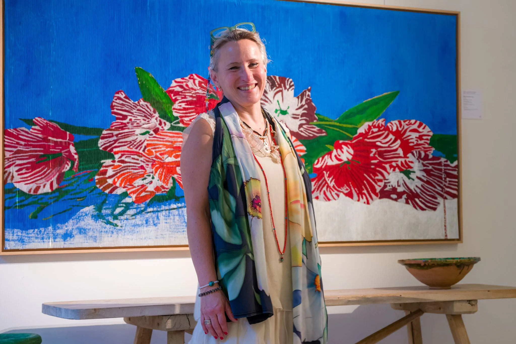 Une femme souriante posant dans une galerie d'art, devant une grande peinture représentant des fleurs rouges et vertes sur un fond bleu, avec une table en bois devant elle.