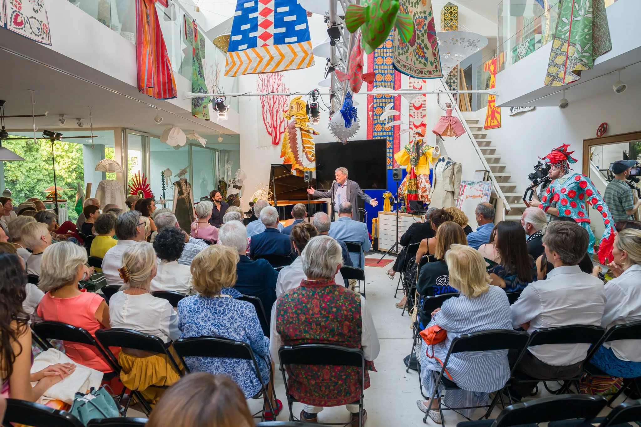 Une présentation ou conférence en intérieur avec un public diversifié et un intervenant au centre, entouré de décors colorés, d'œuvres d'art, de mannequins vêtus de costumes et d'installations artistiques.