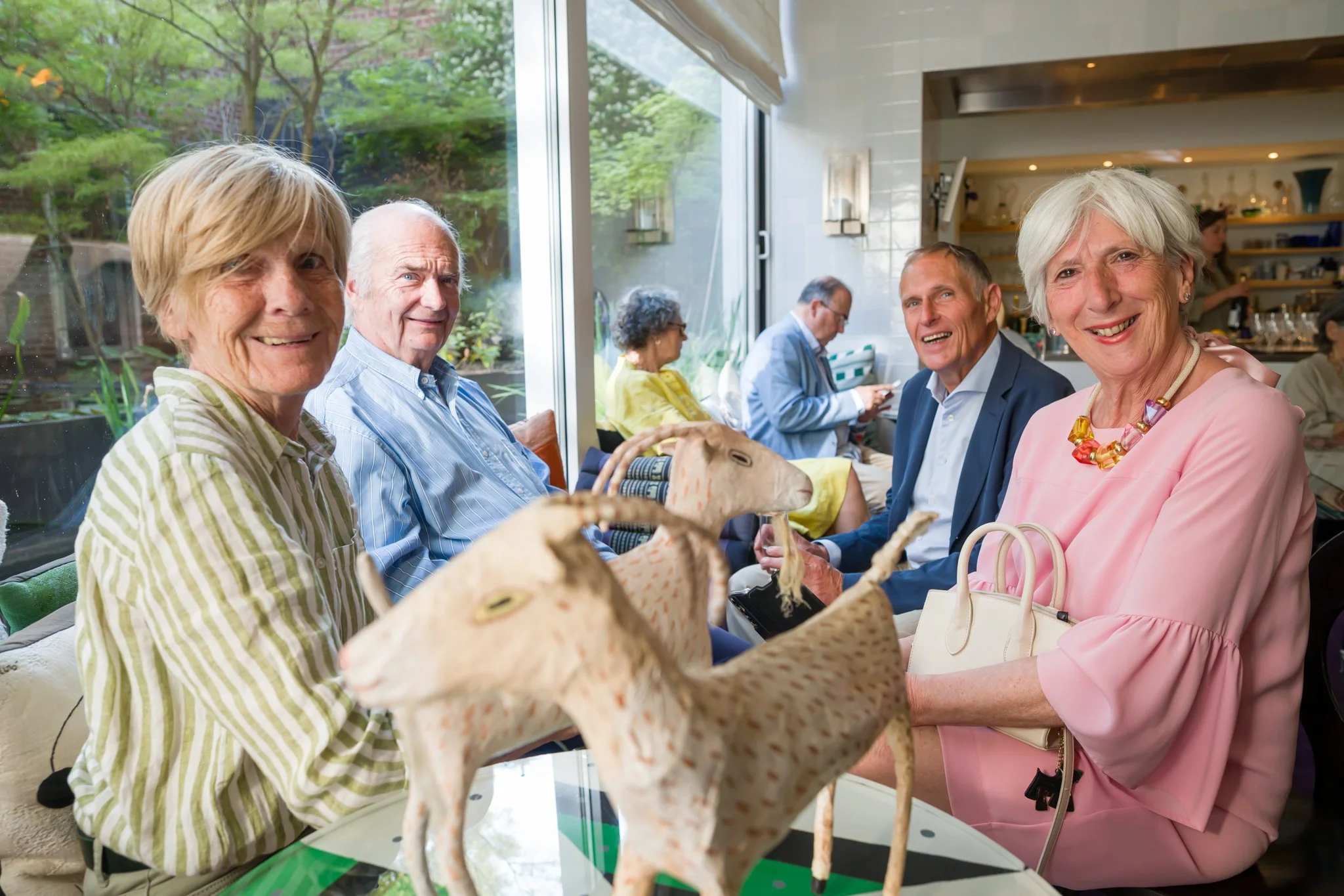 Groupe de personnes âgées assises autour d'une table dans un restaurant, souriantes, avec des poupées en papier sur la table, dans un intérieur lumineux avec fenêtres donnant sur un jardin.