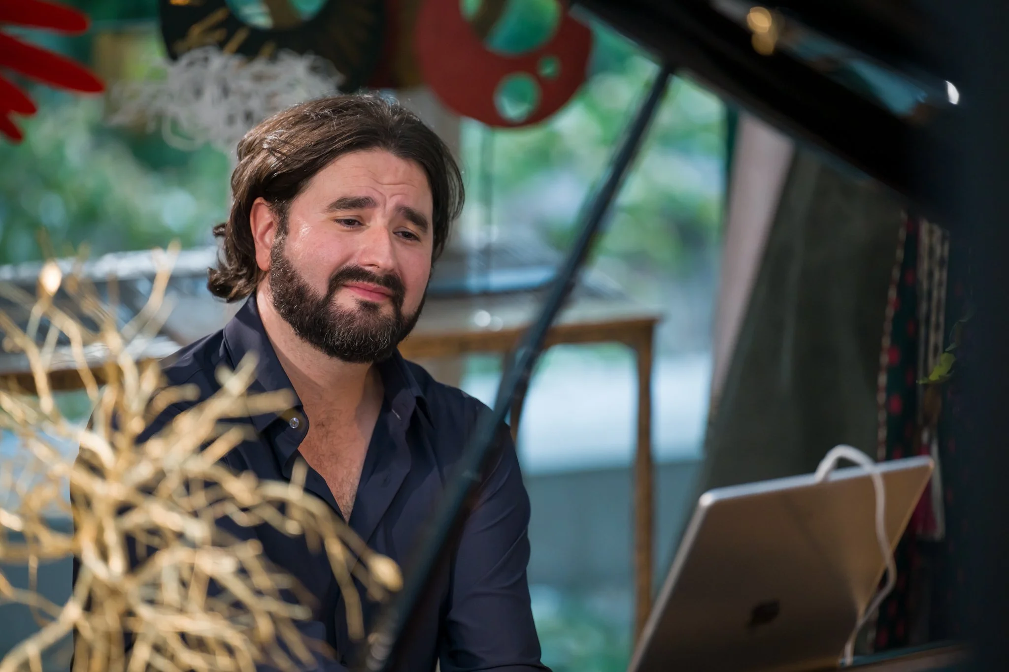 Homme avec barbe et cheveux longs jouant du piano, expression mélancolique, dans une pièce décorée avec des éléments colorés.