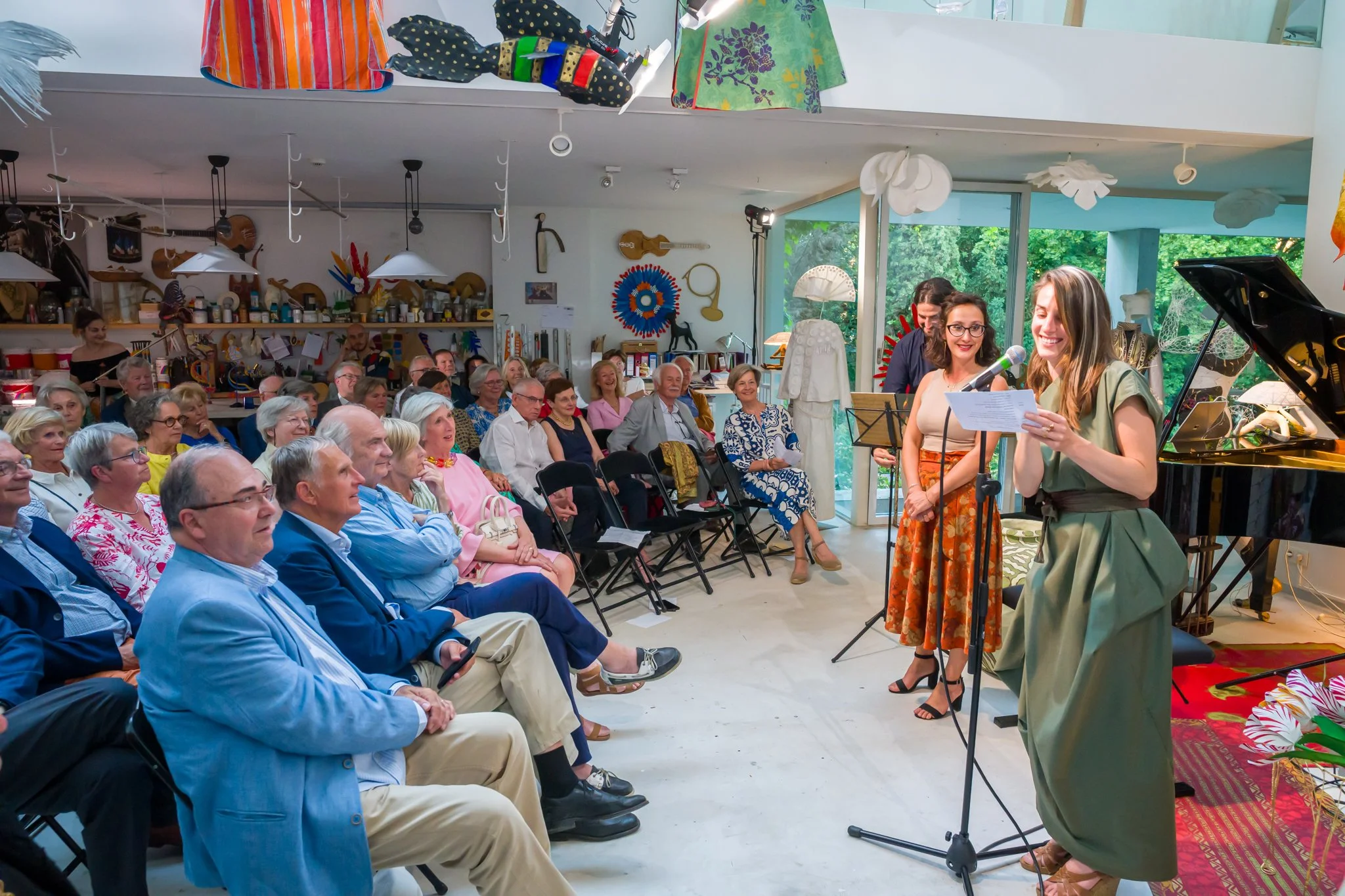 Une femme en robe verte lisant un discours lors d'une performance au piano, entourée de deux autres femmes, au milieu d'un public d'adultes dans un espace intérieur décoré d'objets créatifs et d'artisanat.