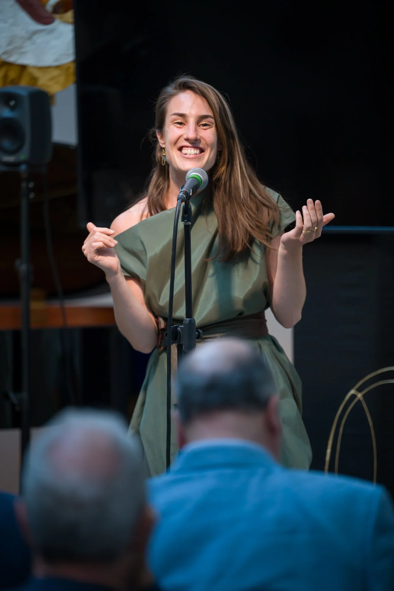 Une femme souriante, avec des cheveux bruns longs, porte une robe verte et parle ou chante devant un microphone lors d'une performance ou d'une présentation, avec un public de personnes âgées en premier plan.