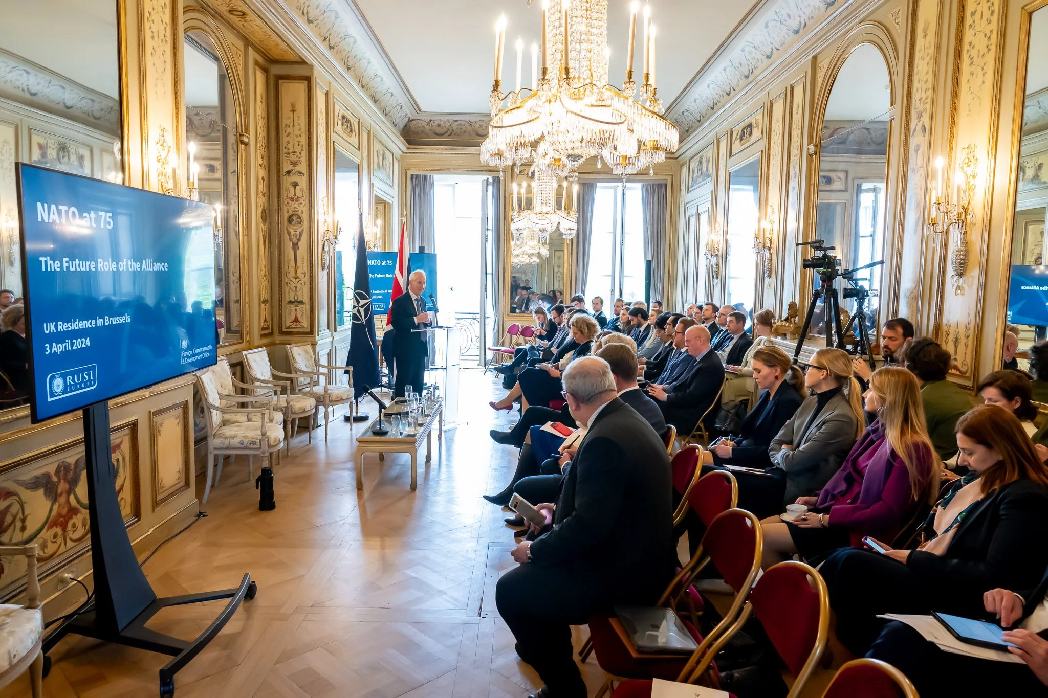 Conference room with seated participants, listening to a presentation on NATO on the occasion of its 75th anniversary, in Brussels. A screen displays details of the event. The room is richly decorated with mirrors and a large chandelier.