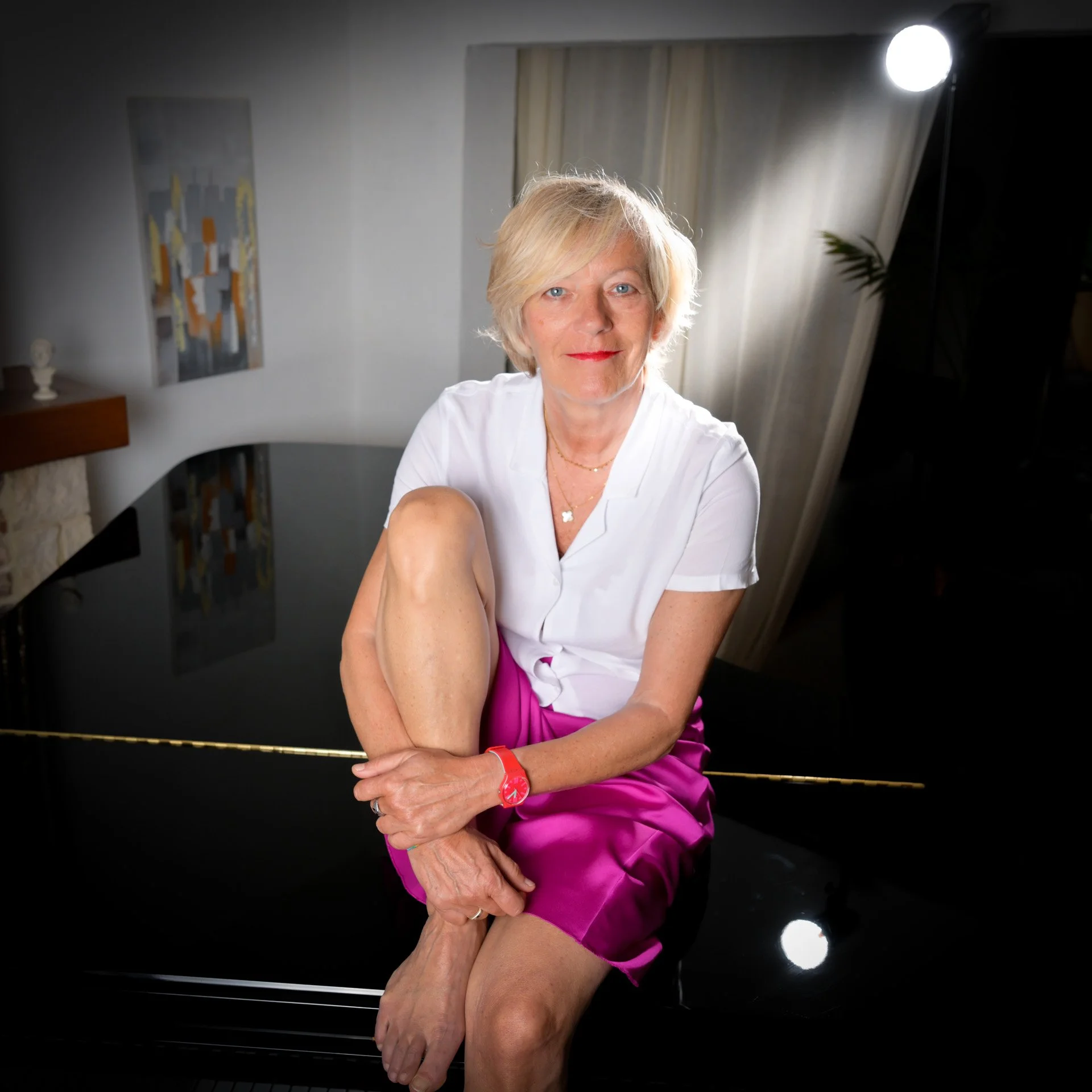 Fabienne Vignier, une femme aux cheveux blonds, habillée d'une chemise blanche et d'une jupe rose, assise sur un piano noir dans une pièce bien éclairée, regardant vers l'appareil photo.