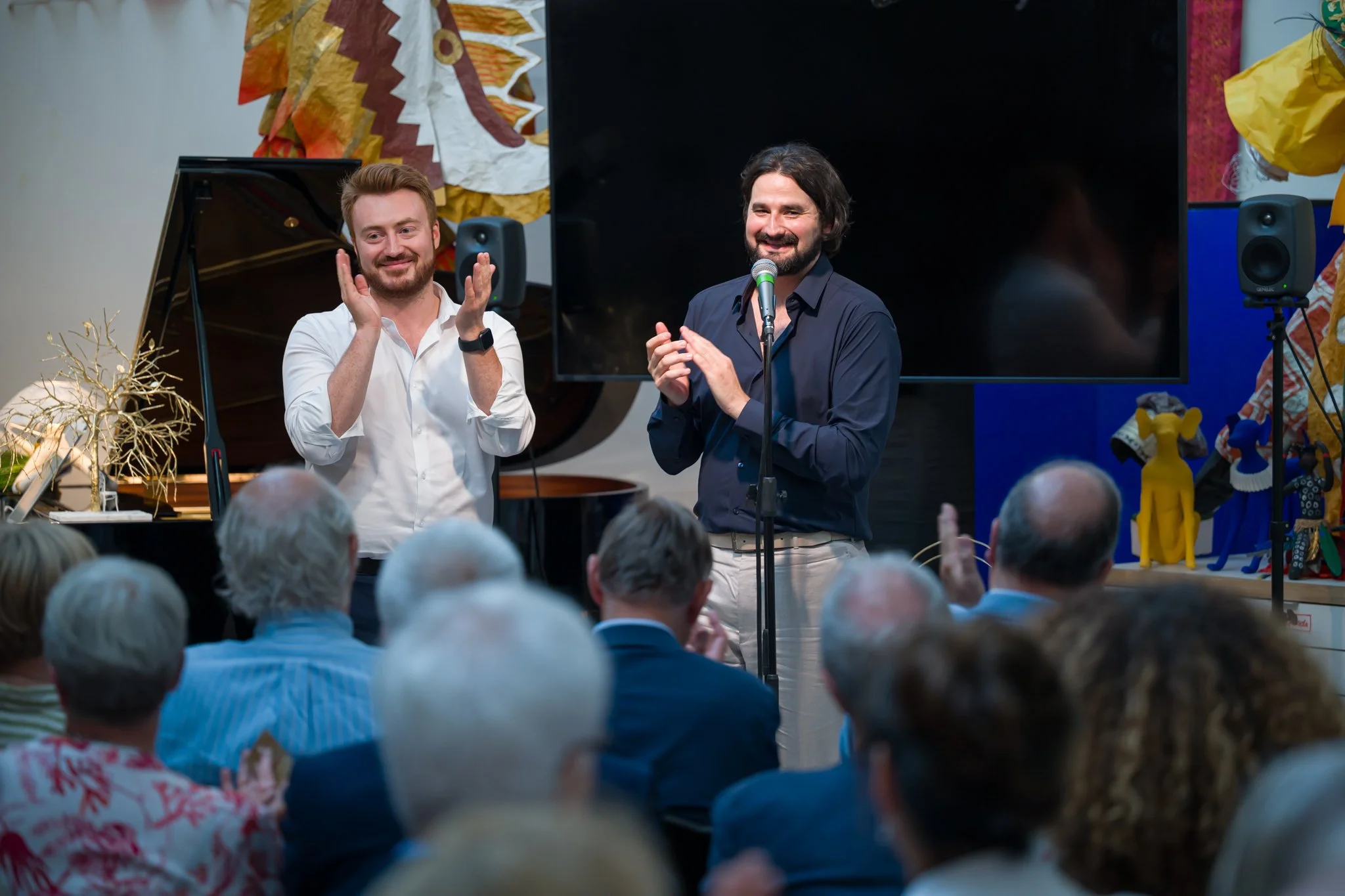 Deux hommes sur scène parlent à un public lors d'un événement culturel, avec un piano à queue noir en arrière-plan, une grande télévision et des décorations colorées.