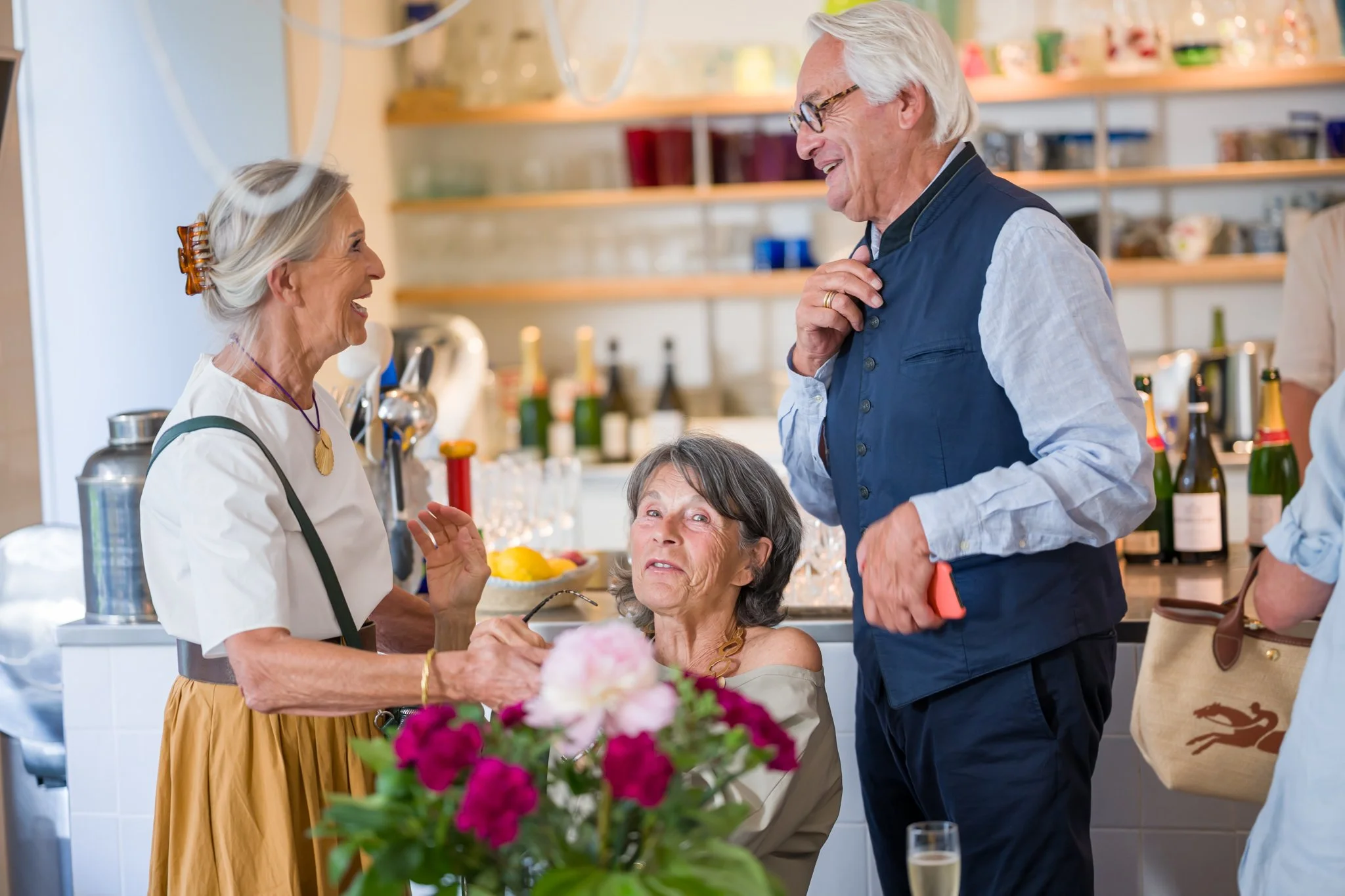Groupe de personnes âgées en conversation amicale dans une cuisine lors d'une fête ou d'un rassemblement, avec des bouteilles de champagne et des fleurs en premier plan.