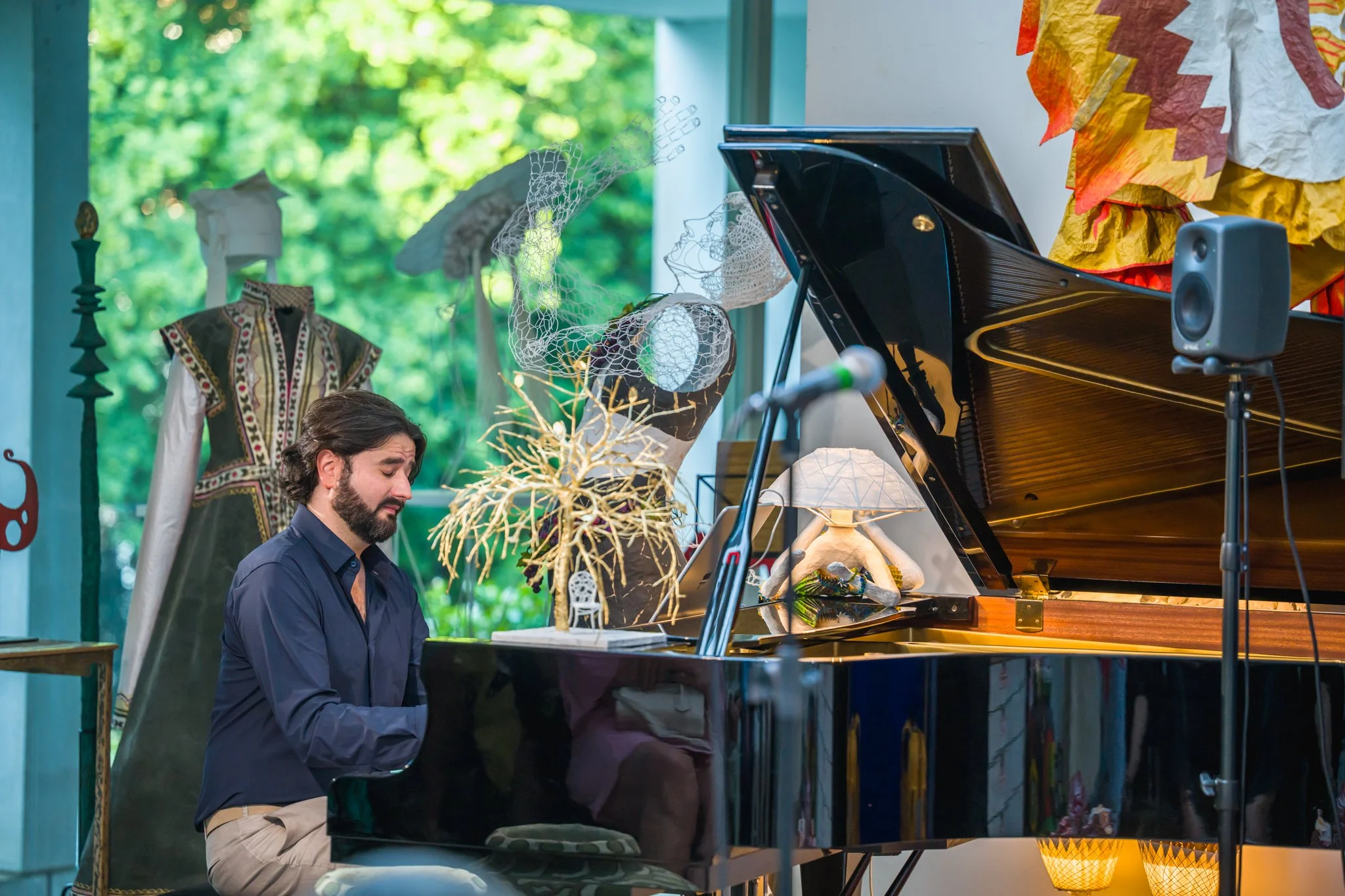 Un homme joue du piano dans un environnement décoré avec des vêtements traditionnels, des lampes artistiques, et un grand piano noir, avec un micro devant lui et une enceinte à côté.