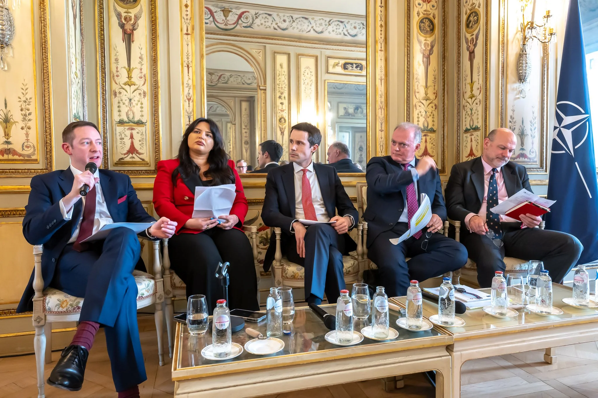 Five people sitting in an elegant lounge, chatting, with documents and bottles of water on a coffee table. A NATO flag is visible on the right. Richly ornate decor and mirror in the background.