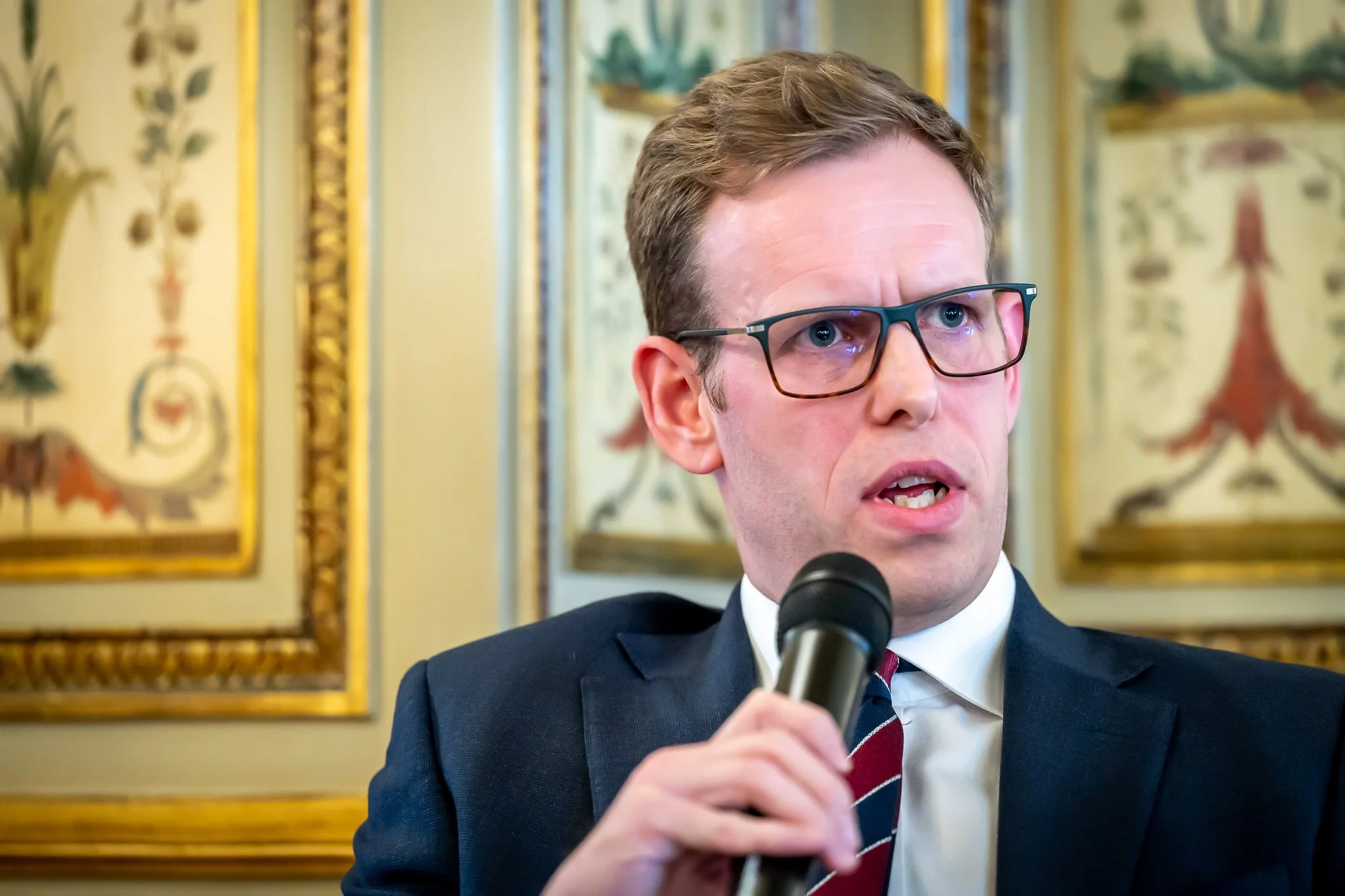 A man in a suit speaks into a microphone, with an elegant interior décor and gold frames in the background.