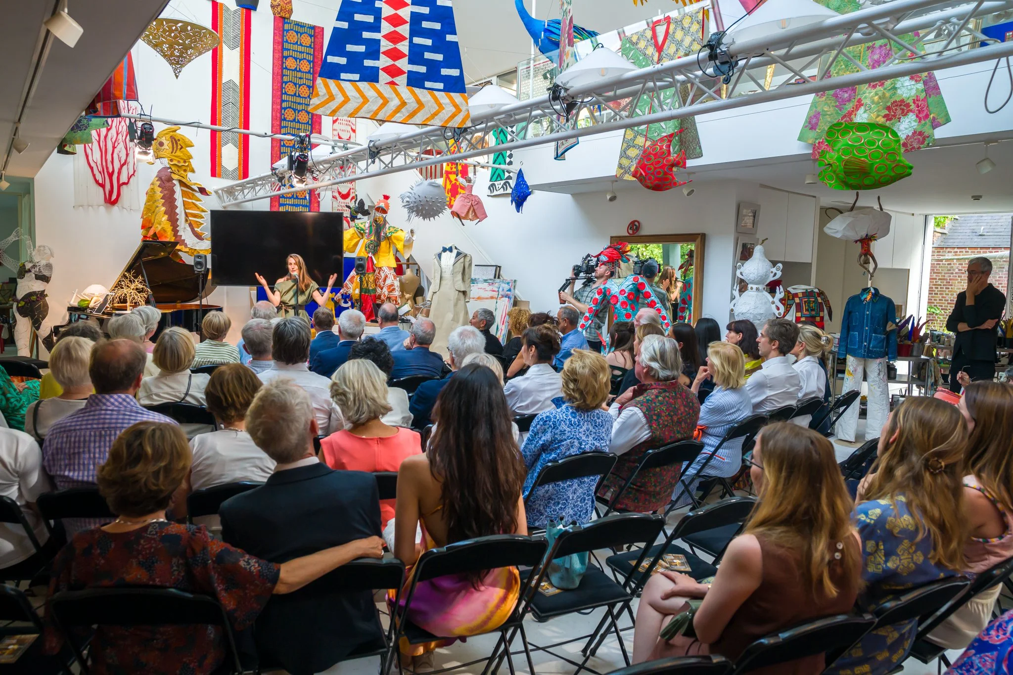 Une foule assise dans un auditorium ou une galerie d'art, regardant une présentation ou une performance artistique. La pièce est décorée avec des œuvres d'art, des mannequins habillés de costumes colorés et des décorations suspendues.