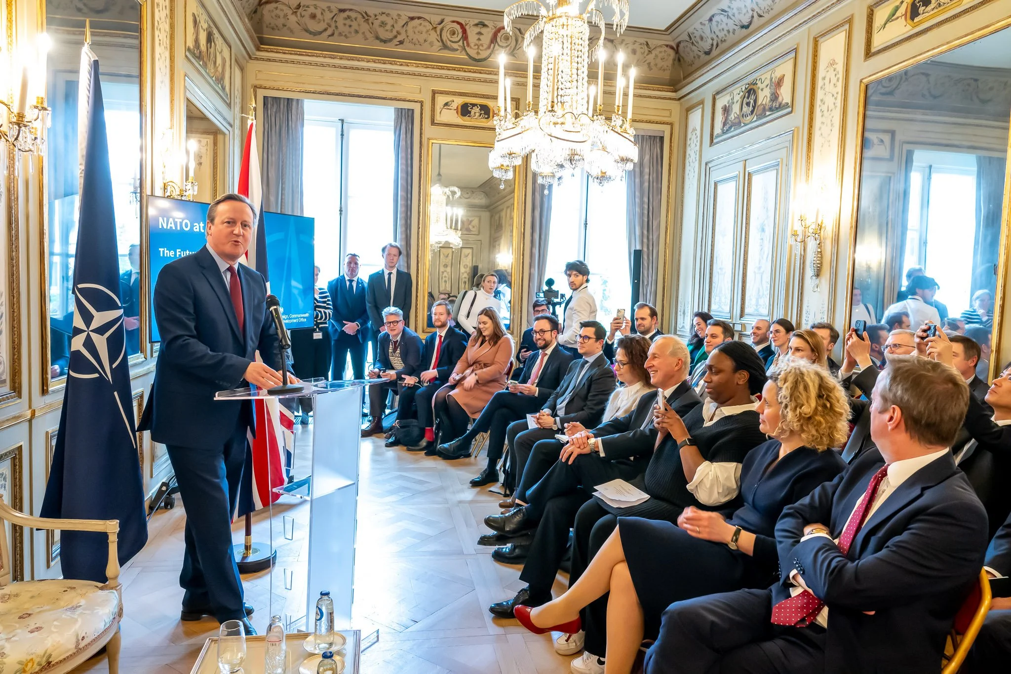 A man in a suit addresses an audience in a room decorated with the NATO flag. A seated audience pays attention, some taking photos. Candelabras and decorative mirrors in the background.