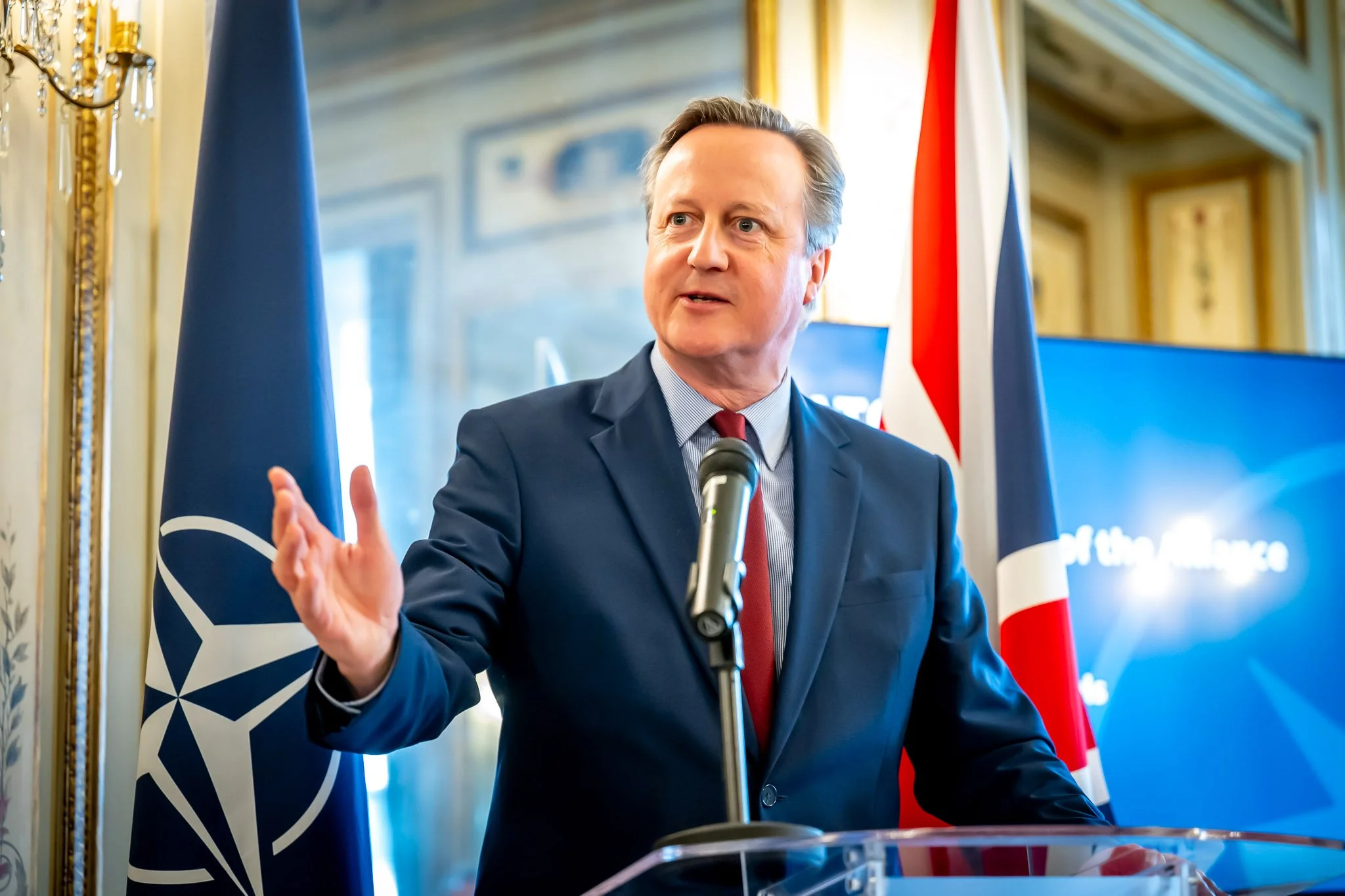 Man in suit speaking at desk with microphone, NATO and UK flags in background.