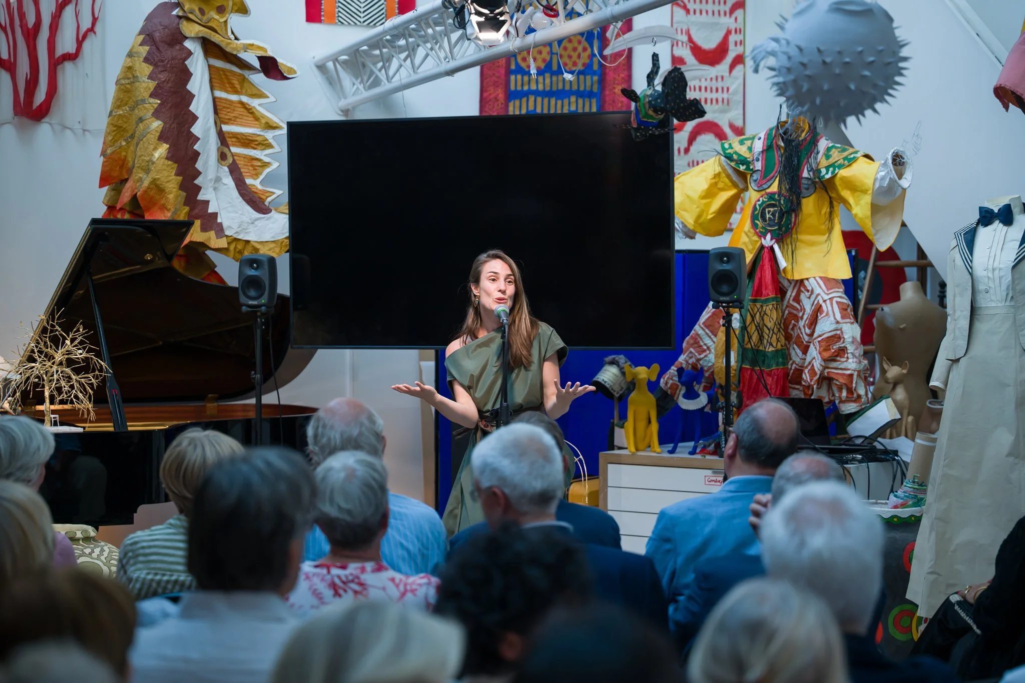 Une femme parle à un micro lors d'une présentation ou d'une performance devant un public. La scène est décorée avec des costumes colorés et des objets artistiques, y compris un piano et un grand écran noir.