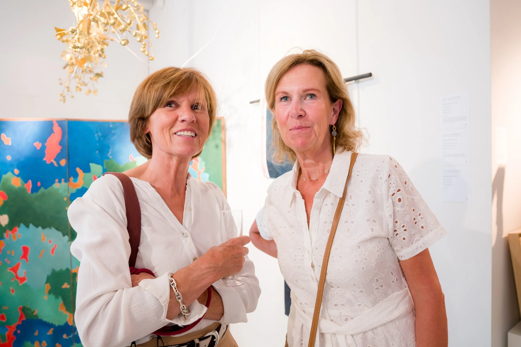 Deux femmes âgées en vêtements blancs à une galerie d'art, souriantes, avec des œuvres d'art colorées en arrière-plan.