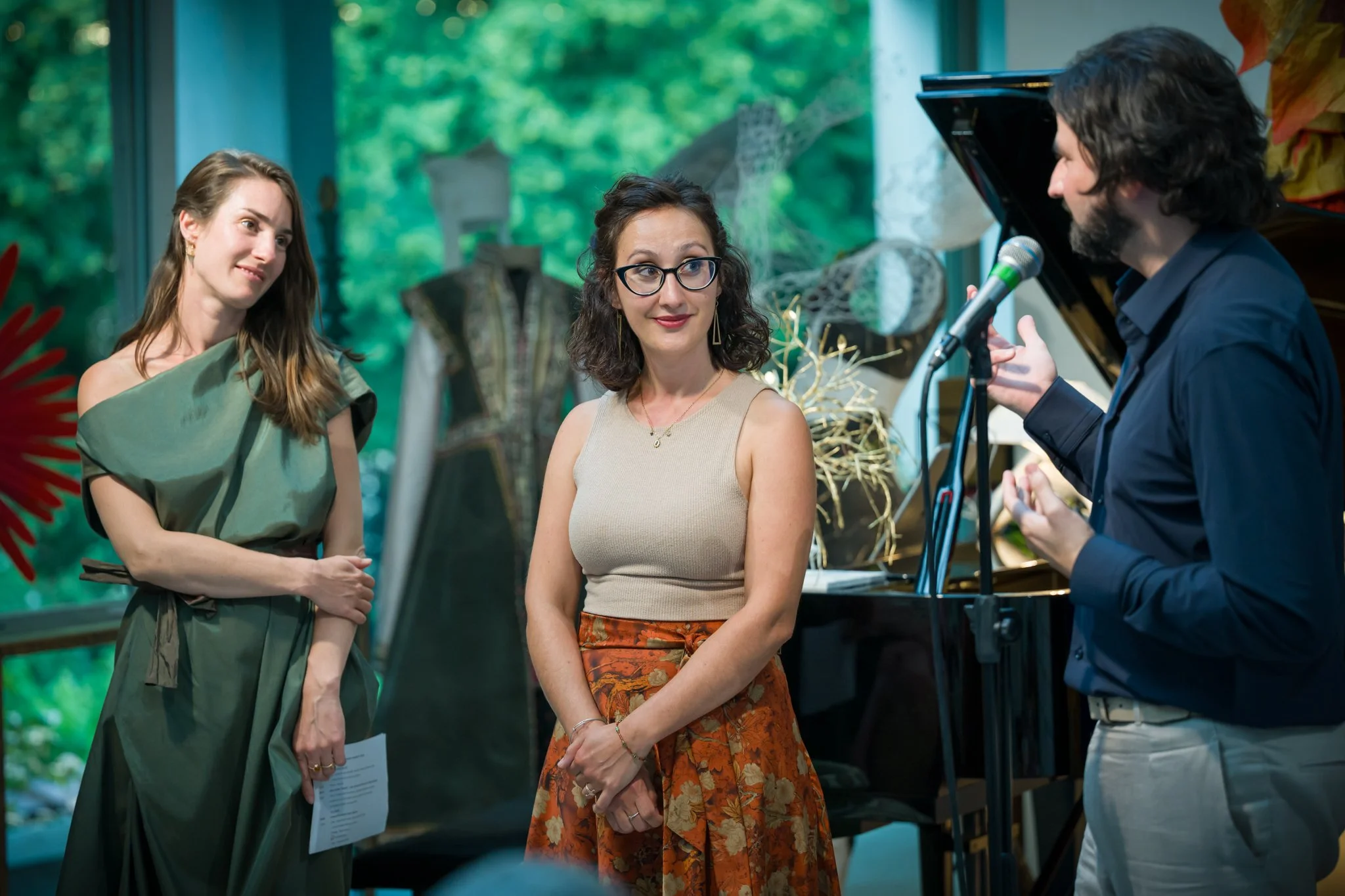 Trois personnes lors d'une conversation dans un intérieur avec décorations artistiques, dont une femme avec des lunettes, une femme avec une robe verte, et un homme avec un piano à queue.
