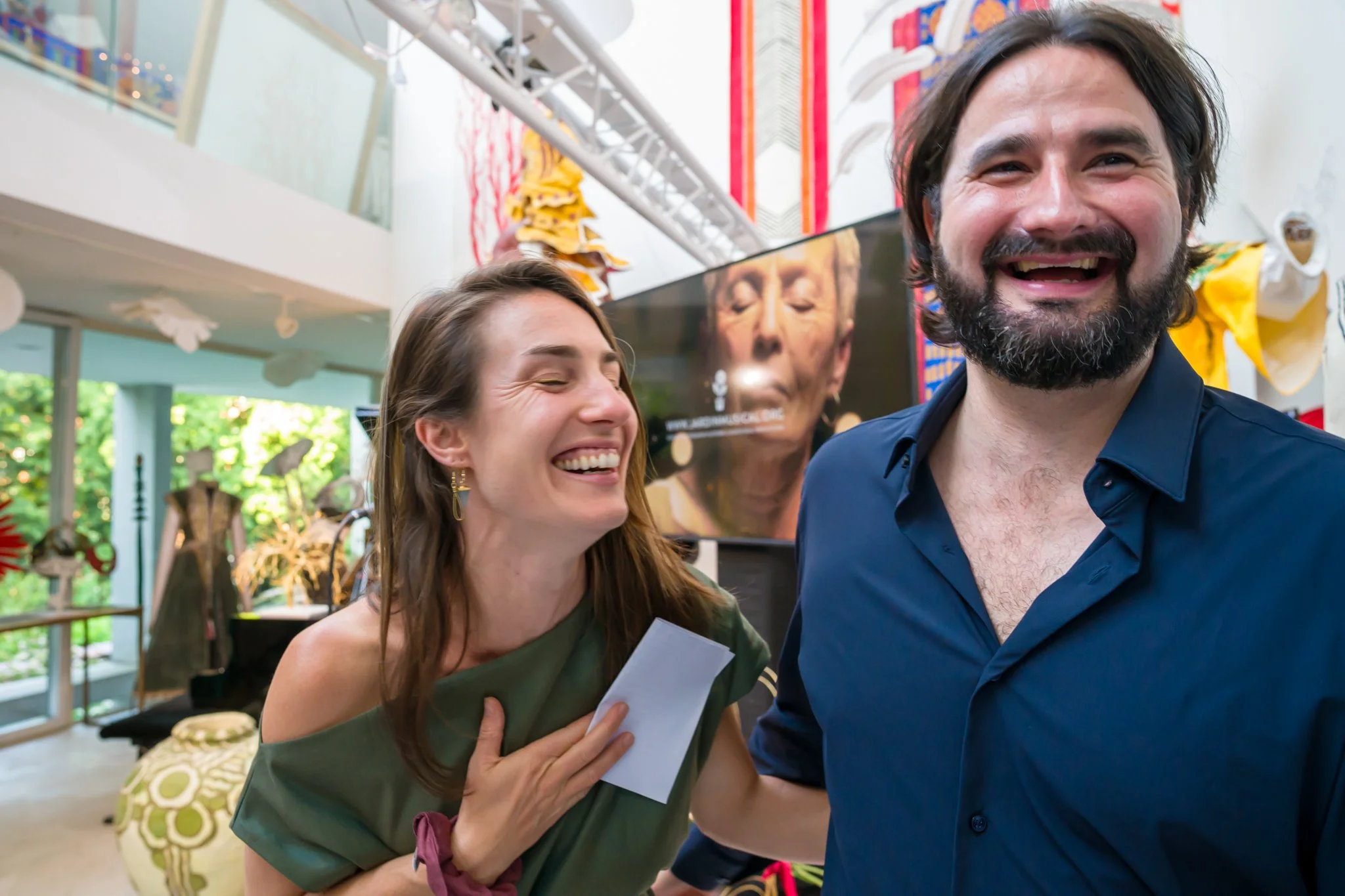 Deux personnes souriantes et riant lors d'une célébration ou d'un événement en intérieur, avec un écran d'affichage derrière elles, dans un espace décoré avec des œuvres d'art et des objets colorés.