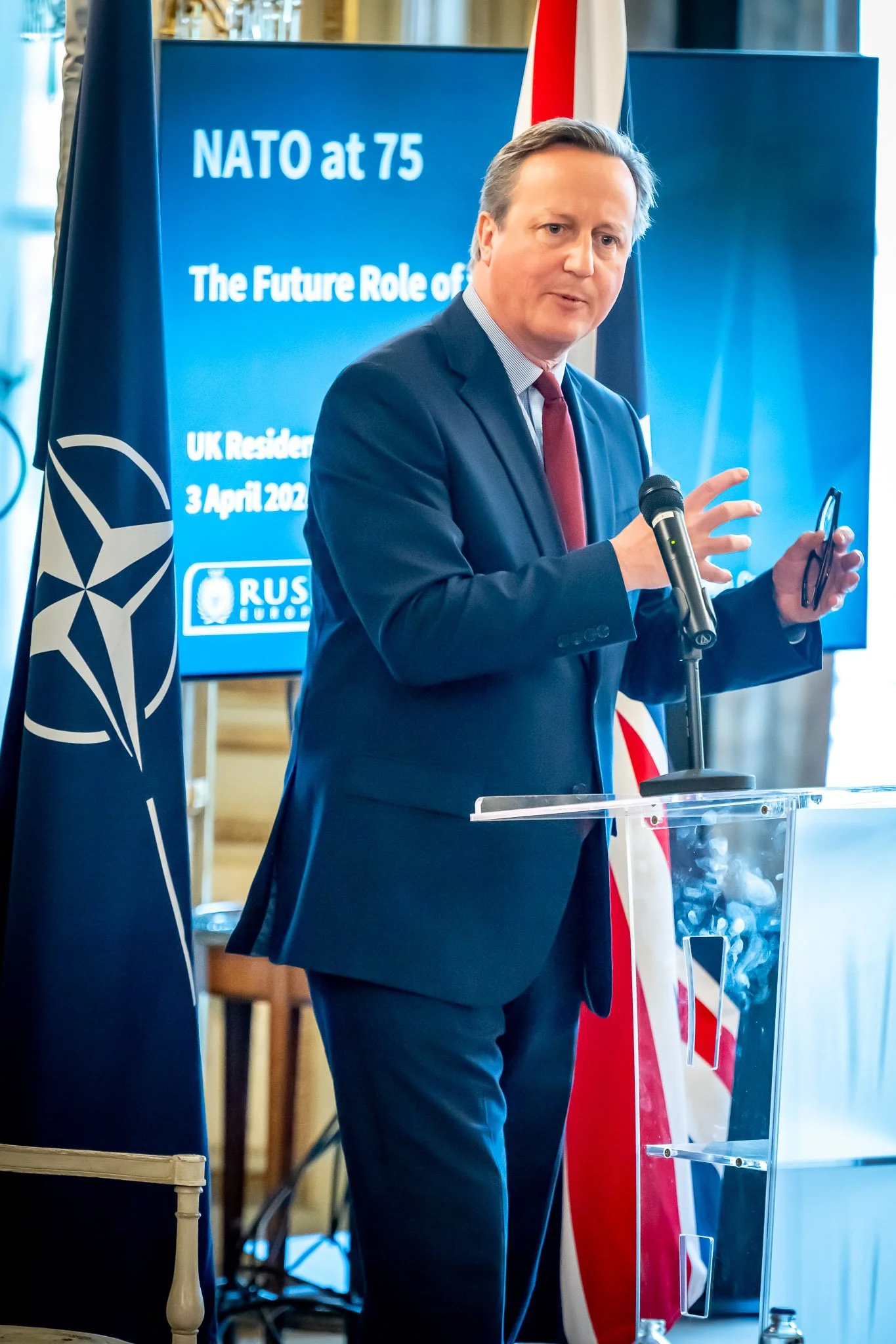 A man in a suit speaks at a lectern with a screen and a flag behind him. The screen reads "NATO at 75". A microphone is attached to the lectern and a NATO flag is visible next to it.