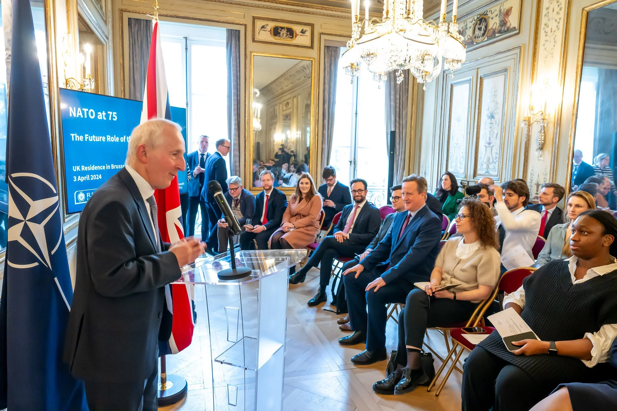 A conference is taking place in an elegant room, with a speaker at the microphone. A screen displays "NATO at 75" and "The Future Role of the UK". Participants sit and listen attentively. Flags and mirrors adorn the room.