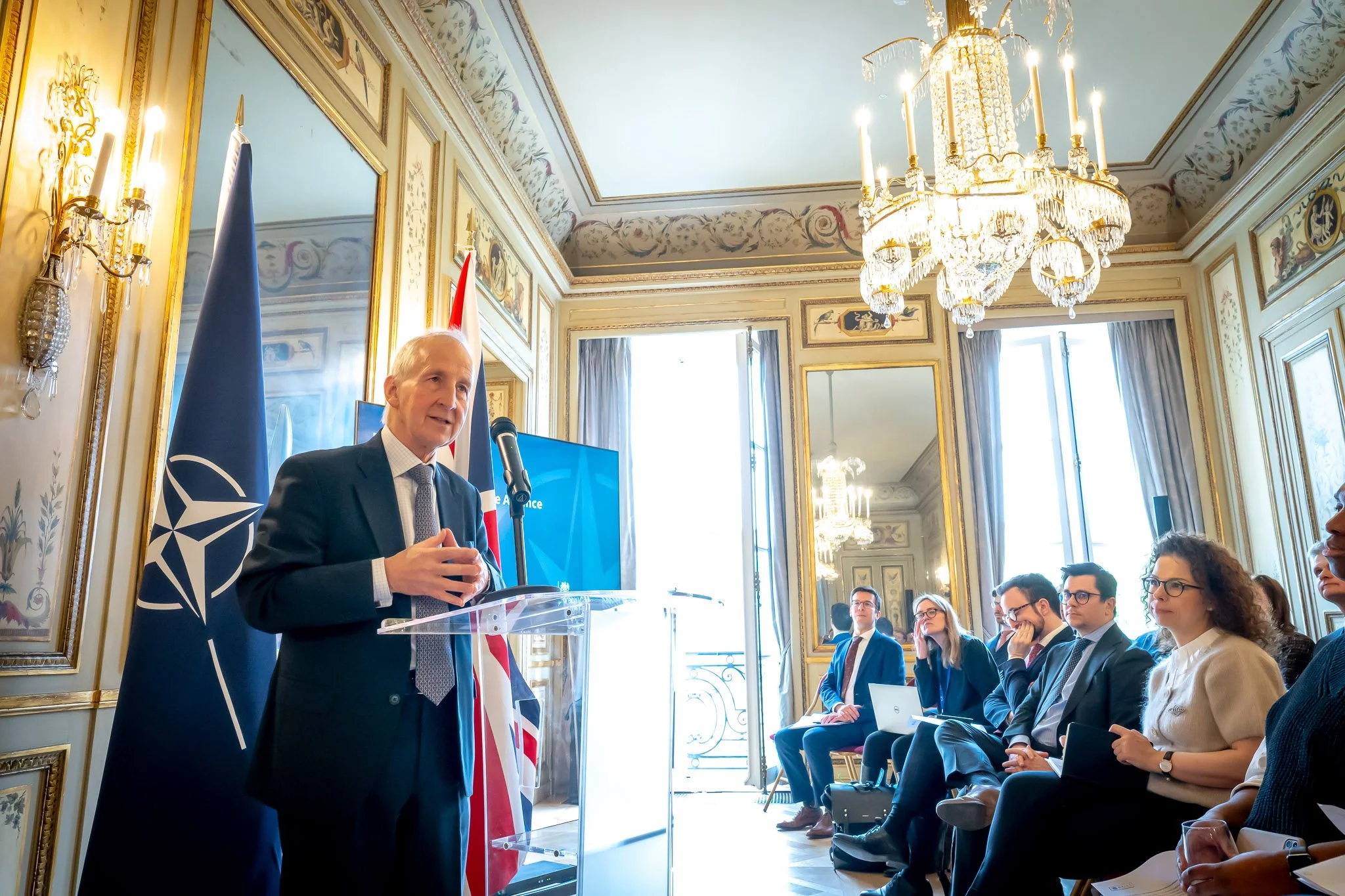 A man in a suit speaks into a microphone at a conference, with NATO and UK flags in the background. Seated people listen attentively in an elegant room with chandeliers and mirrors.