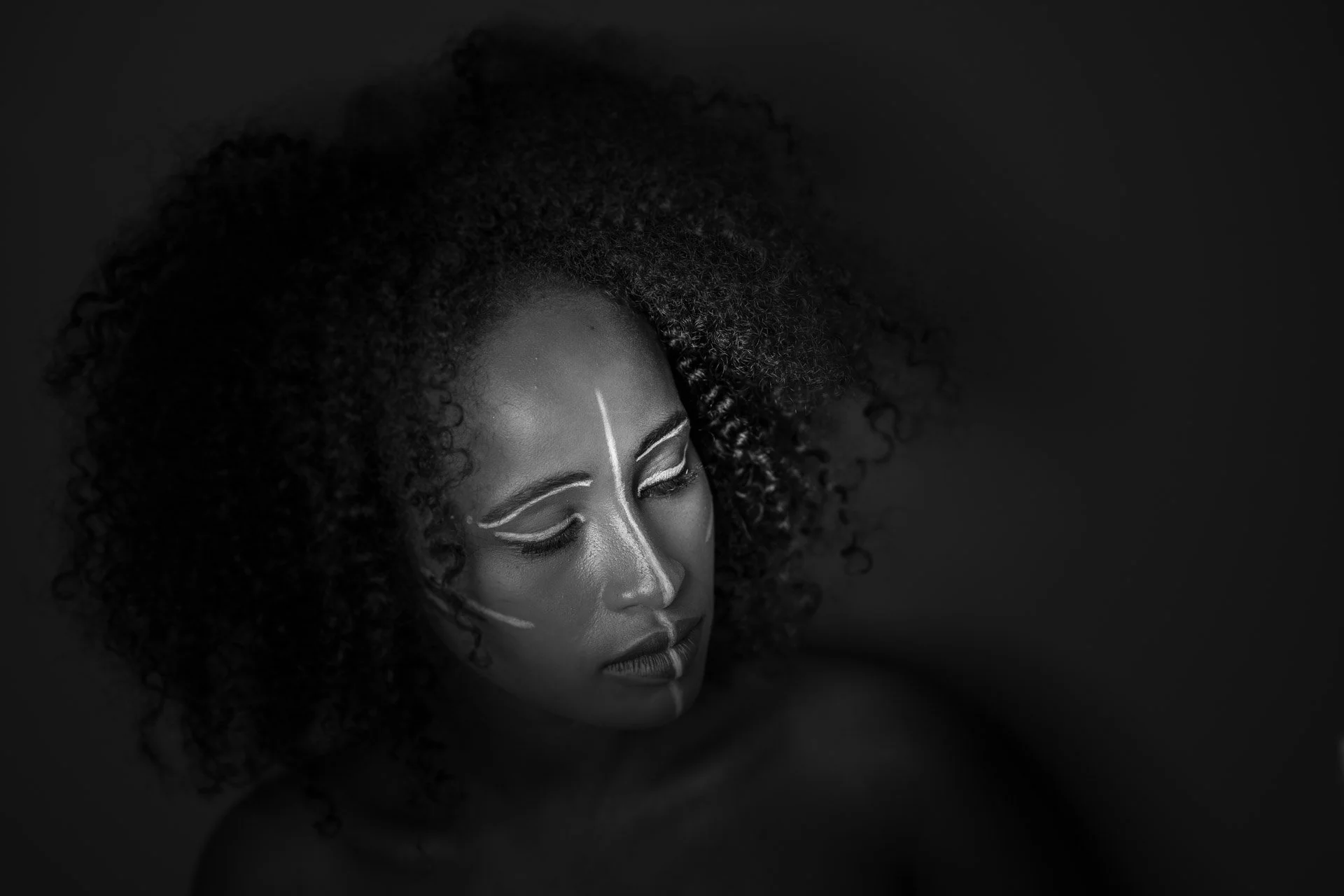Portrait en noir et blanc d'une femme avec des cheveux bouclés, portant du maquillage artistique blanc sur le visage, avec les yeux fermés et une expression sereine.