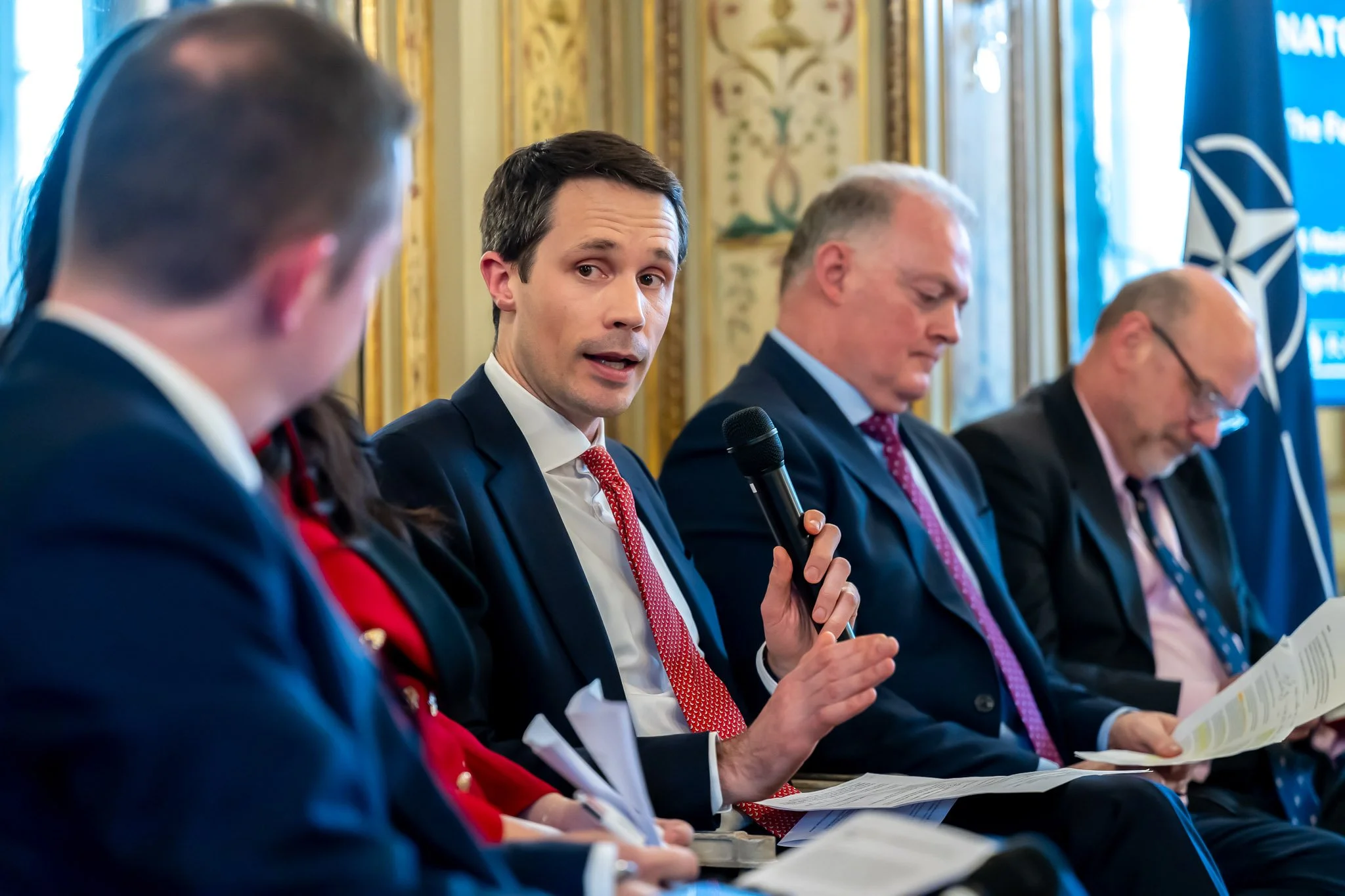 A group of people in formal costumes are taking part in a discussion or conference. One of them holds a microphone and speaks, while others listen or take notes. A NATO flag is visible in the background.