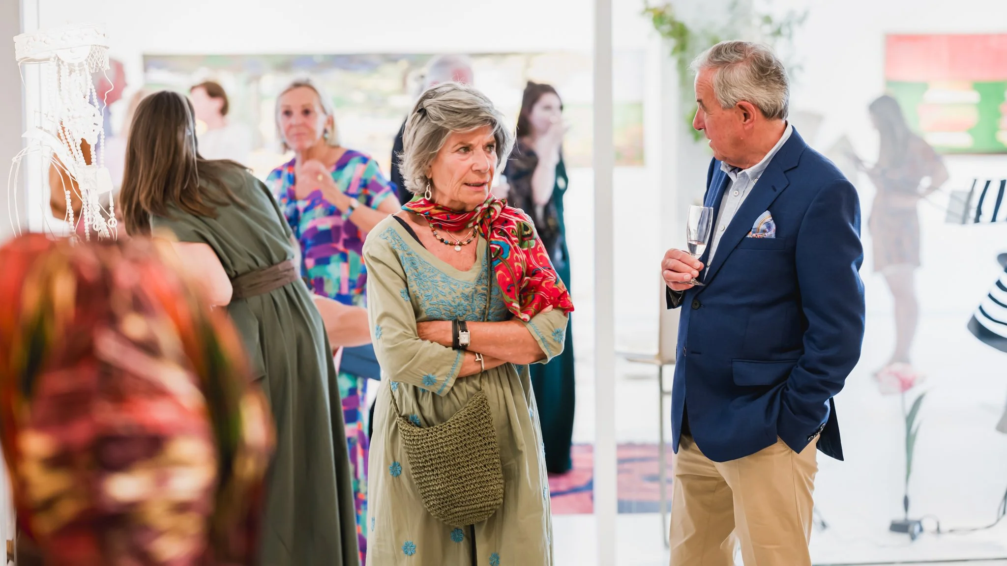 Deux personnes âgées discutent lors d'une fête avec plusieurs invités en arrière-plan, à l'intérieur, avec une ambiance festive.