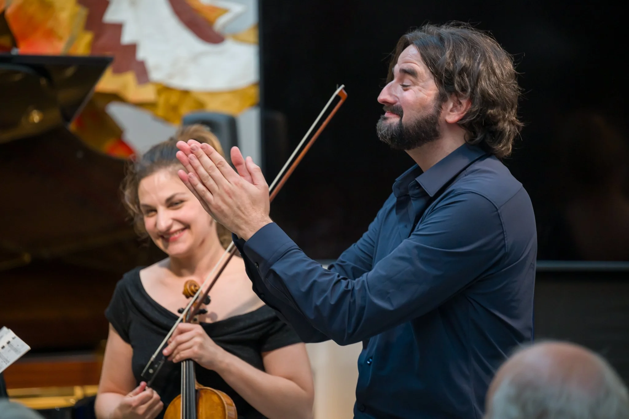 Un homme et une femme un groupe de musiciens en concert, l'homme semble remercier le public avec un sourire, la femme tient un violon, tous deux joyeux.