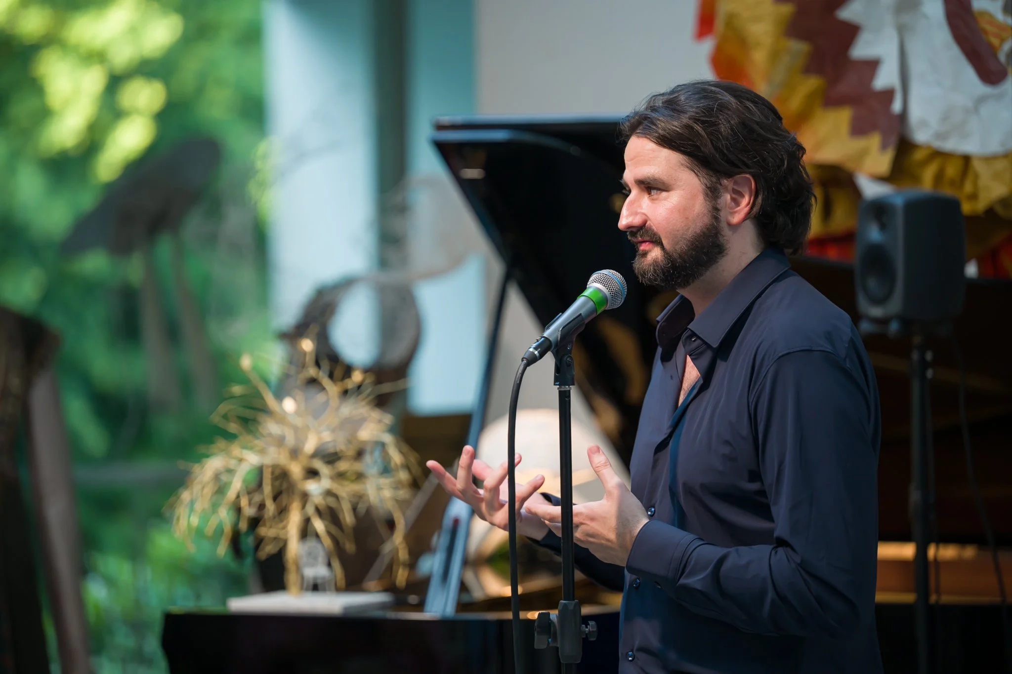 Un homme avec une barbe et des cheveux longs, parlant dans un microphone lors d'une performance, avec un piano à queue en arrière-plan et une décoration de Noël.